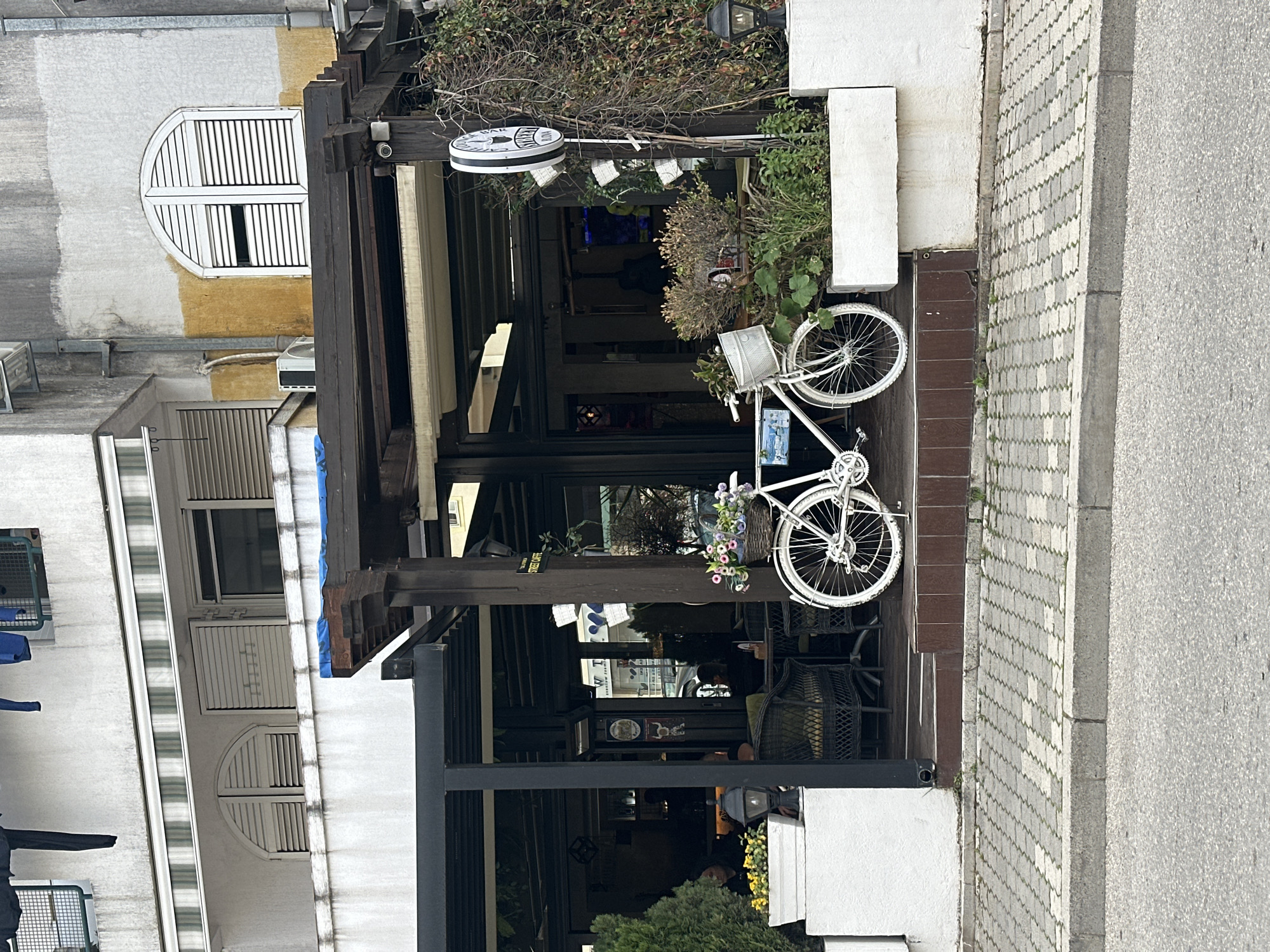 The image captures a street scene in Budva, Montenegro, featuring a restaurant or cafe. A white decorative bicycle adorned with flowers is mounted on the front facade. The restaurant has an outdoor seating area with dark wicker chairs visible inside. The architecture consists of a building with white walls and brown wooden accents, including window frames. There is a partially visible sign with the words "STREET CAFE". The street outside is paved with interlocking concrete blocks. The photo is taken at eye level.