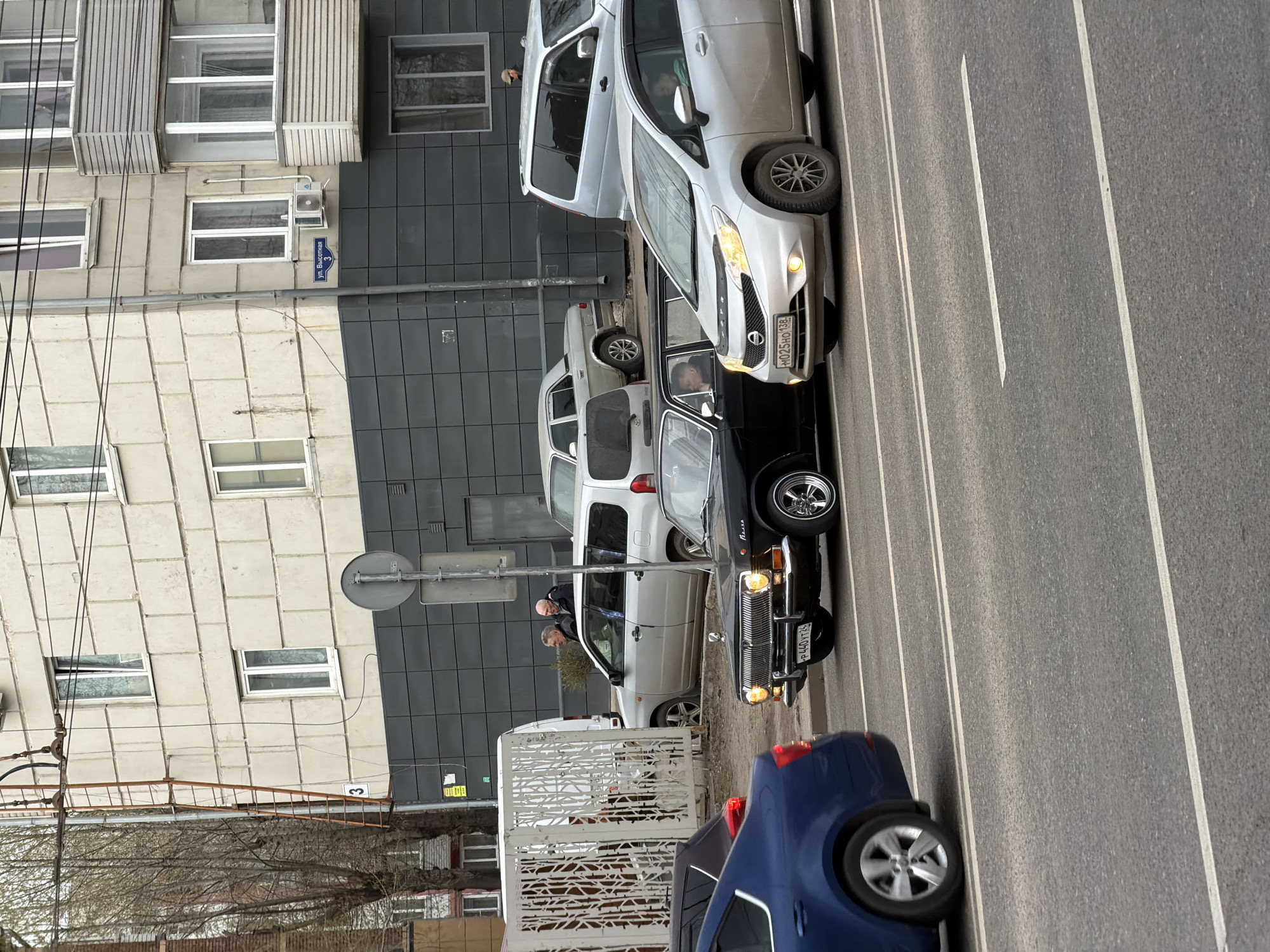 The image shows a street scene in Krasnoyarsk, Russia. A black Volga automobile with license plate Я440ТТ54 is parked on the street, adjacent to a white Toyota Probox. Further up the street, a silver Nissan Note, bearing license plate Н025НО138 is also parked. A dark-colored car is visible at the bottom of the picture. Two men stand on the sidewalk next to the Toyota. A building with beige tiled walls and a dark gray facade is in the background, marked with the street address "ул. Высотная 3". The street has white lane markings. The photo is taken from an elevated angle.