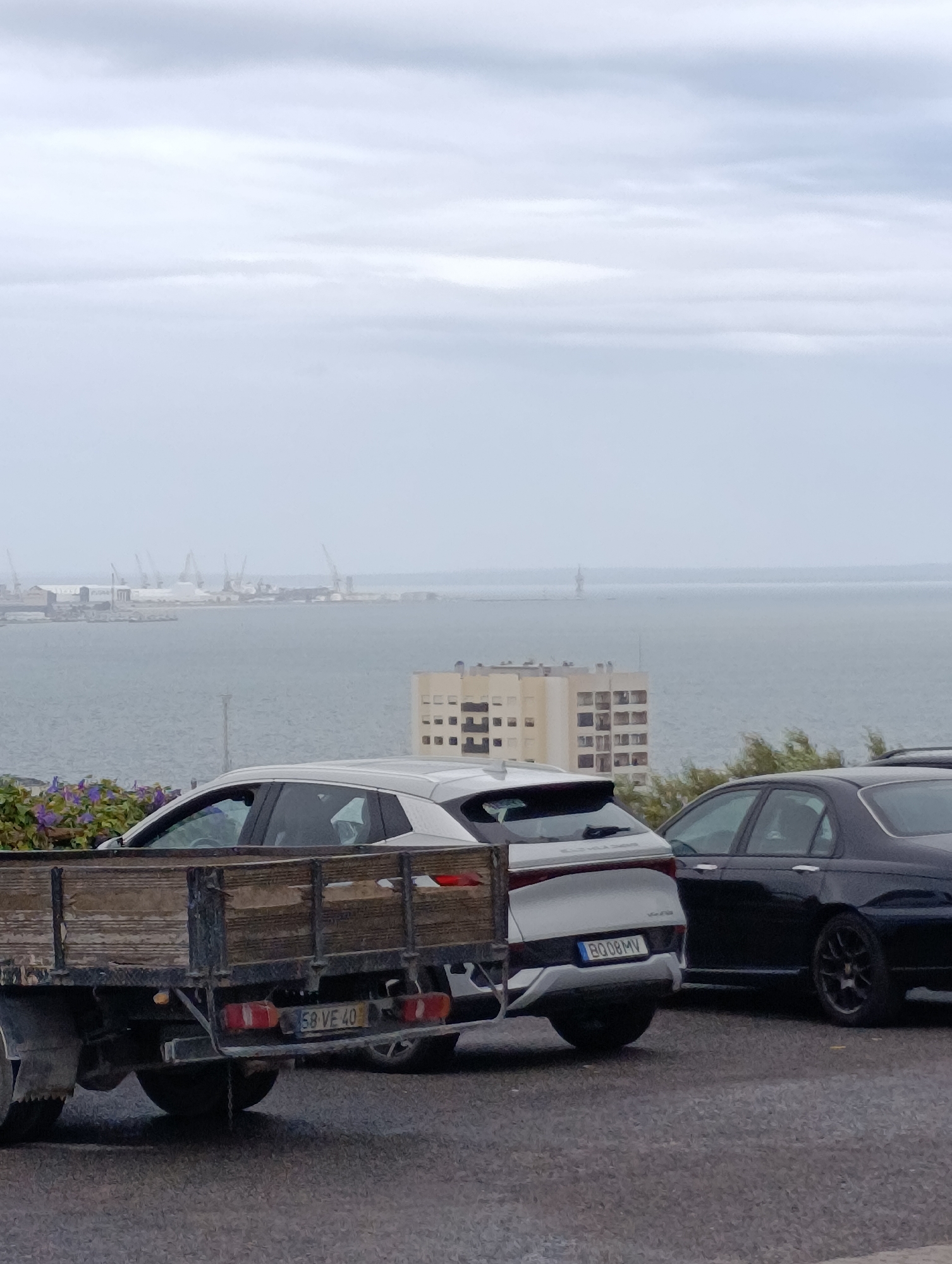 The image depicts an outdoor scene at what appears to be a port or industrial area in Setúbal, Portugal. In the foreground, a weathered wooden truck bed is visible, with a license plate reading "58 VE 40". To the right of the truck, a silver SUV is parked, and further to the right is a black sedan. Beyond the vehicles, a large body of water is present, with a distant industrial waterfront featuring cranes and buildings. In the mid-ground, a multi-story building with a distinctive grid-like facade stands out. The sky is overcast, suggesting a cloudy day. The overall impression is one of a working port environment under a muted sky.