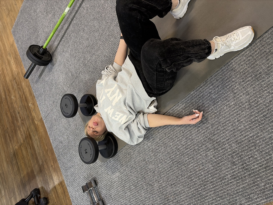 The scene depicts an indoor fitness facility, specifically a gym or workout area, located in Chaikovsky, Russia. A light-skinned female with blonde hair is lying supine on a dark grey exercise mat, centered in the frame. She wears a light grey hooded sweatshirt with "NEW YORK" and "NY" printed in white, dark grey acid-wash sweatpants, and light-colored athletic sneakers (Nike brand visible). A dark smartwatch is on her left wrist, and her fingernails are painted light pink. Her mouth is open with her tongue extended, indicative of fatigue or a playful gesture. Two black Technogym dumbbells, each marked "14," are positioned on the mat near her head, one on either side. To the left of the mat, a stack of chrome dumbbells is partially visible on the floor. To the right, a green and black barbell with a single weight plate rests on the floor. The mat and equipment are placed on a light grey, ribbed carpet. Beyond the carpet, a section of light brown wooden flooring is visible. The lighting is even, illuminating the entire area.