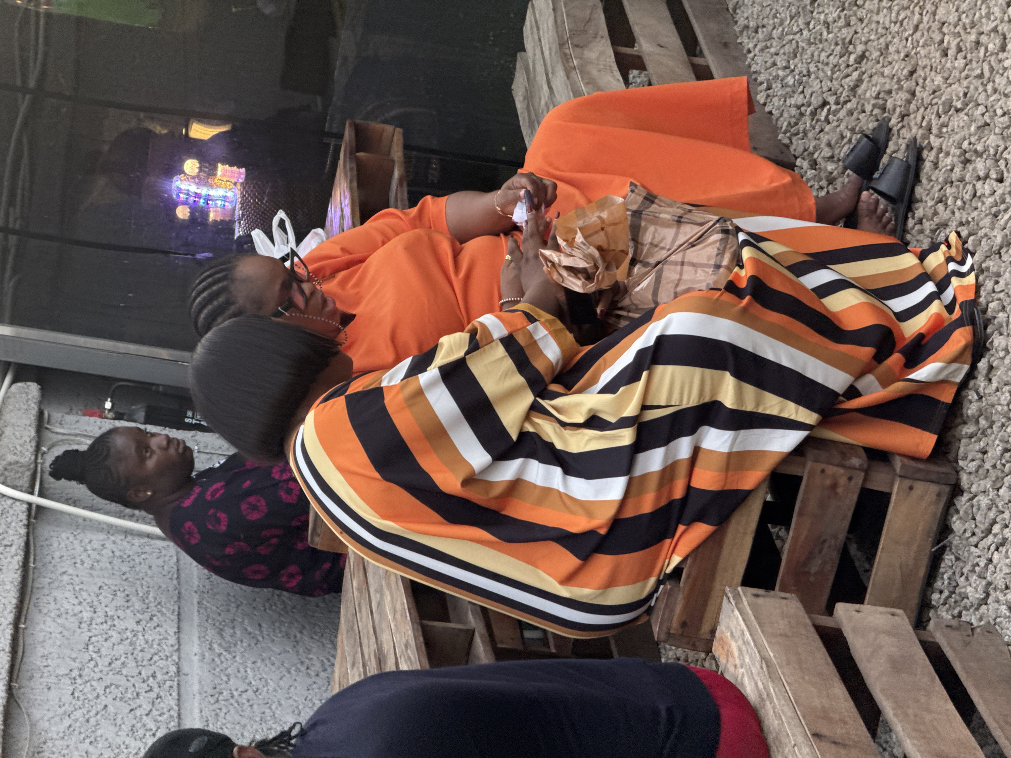 The image depicts four individuals in an outdoor setting in Gwarinpa, Nigeria. They are seated on wooden pallets that serve as furniture. One woman wears an orange dress, and another has on a striped dress in shades of orange, yellow, black, and white. She is holding a crumpled brown paper bag and a white tissue in her hands. A third woman is visible, wearing a black top with a red patterned design. Another person wears a dark blue shirt. The ground is covered in gravel. A wall or panel is visible behind the group, with a neon light display reflected on its surface.