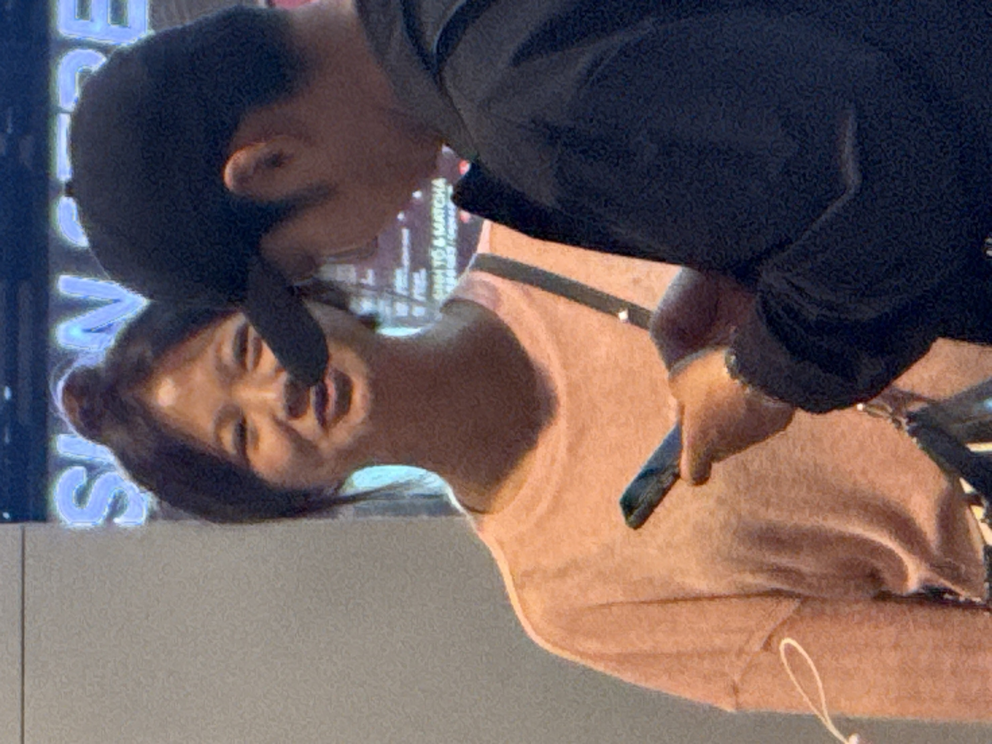 In Bắc Cam Ranh, Vietnam, a young Asian woman with her hair tied in a bun is looking forward and smiling. She is wearing a light peach-colored shirt. Beside her, a man wearing a black baseball cap and a dark jacket is holding a smartphone, with his face obscured by the cap. They appear to be in a public space, possibly a mall or entertainment venue, indicated by the neon sign in the background with blue lettering. The woman has a small white earbud cord hanging from her left ear. A dark bag with a pattern is visible on her right shoulder.