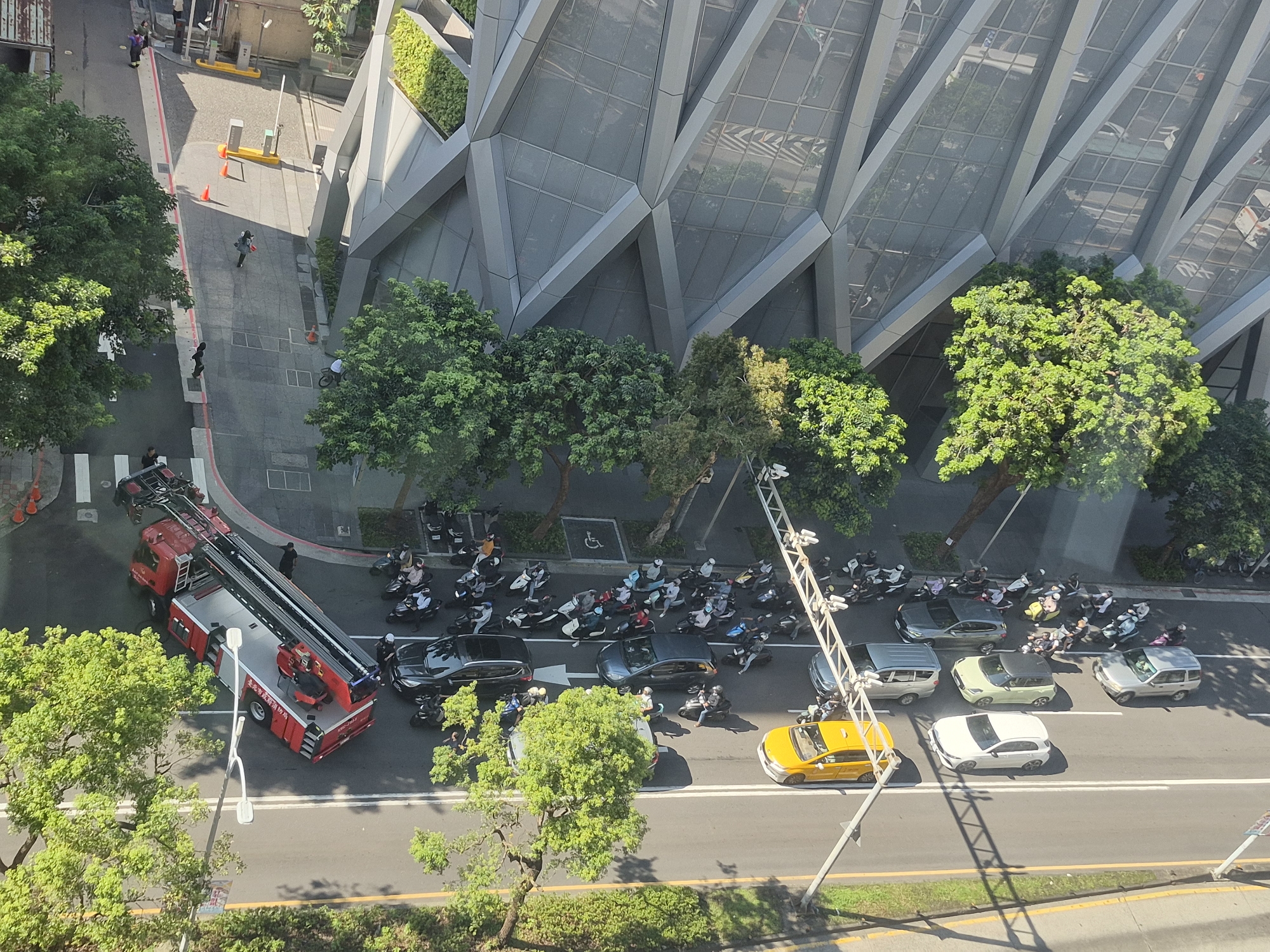 The image captures a street scene in Taipei, Taiwan, looking down from a high vantage point. A large red fire truck with an extended ladder is parked on the road, facing right. Several people are standing around the fire truck. On the road, a line of vehicles, including cars and numerous scooters, is visible. A yellow taxi is among the vehicles. A modern building with a distinctive angular, glass facade stands to the right of the street, with trees in front of it. The road is asphalt with lane markings. The environment appears to be a sunny day.