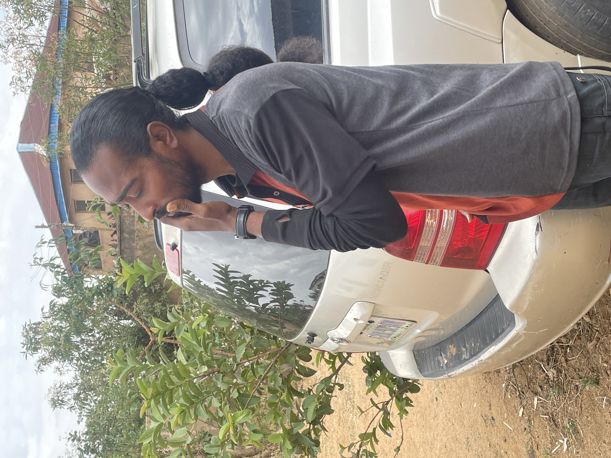 A man with dark hair pulled back in a ponytail stands close to the rear of a light-colored Toyota Highlander. He is holding a small dark object near his mouth. The car has a license plate partially visible, and the word "HIGHLANDER" is visible on the rear. The environment includes greenery and a building in the background. The location is likely in Jos, Nigeria.