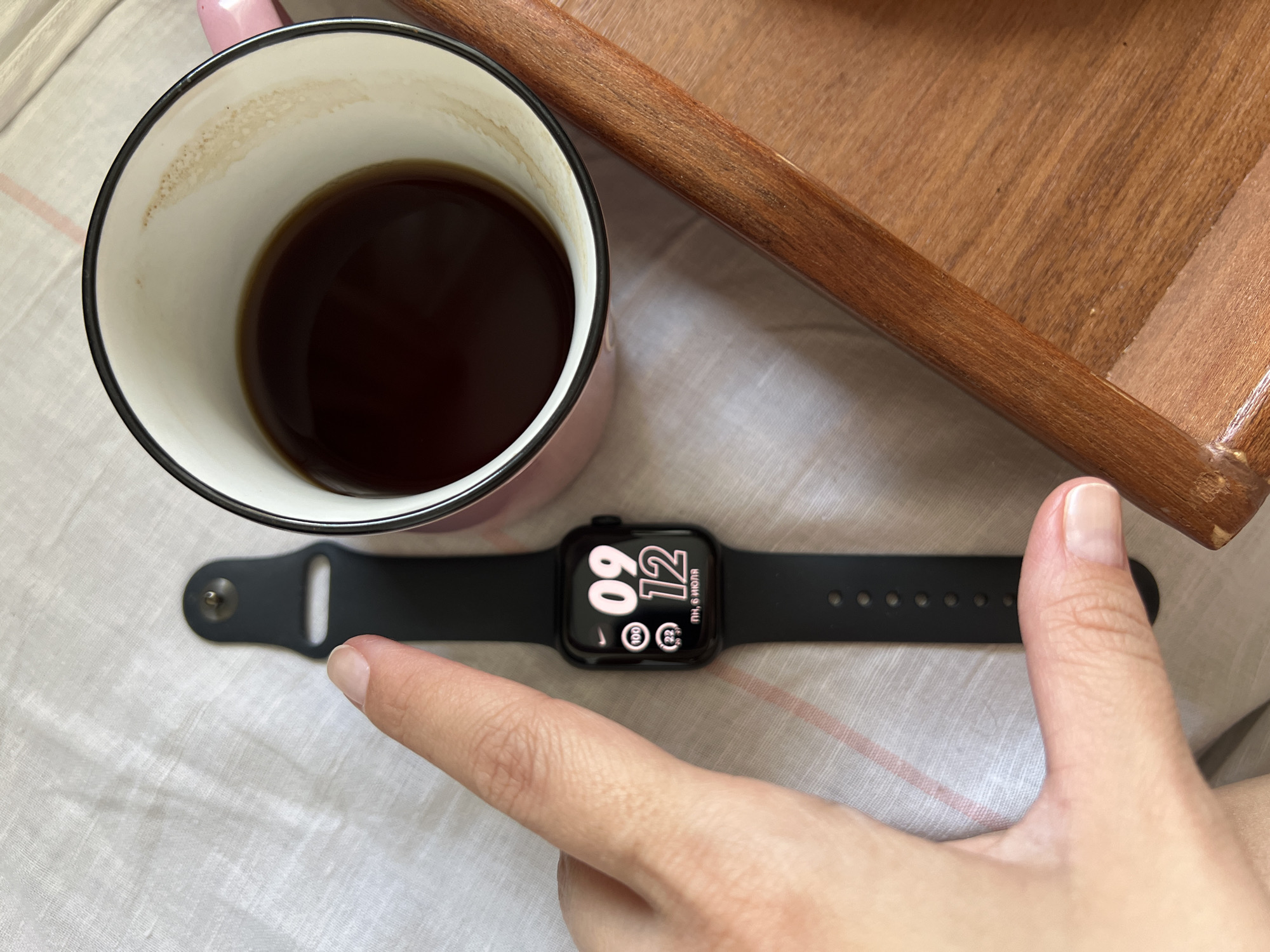 A close-up, top-down shot shows a hand with a light skin tone and light-colored fingernails, partially obscuring a black Apple Watch. The watch face displays the time as 09:12, along with icons for "100" and "22," and the text "ПН, 6 ИЮЛЯ." To the right of the watch is a pink mug with a black rim, filled with dark coffee, showing some residue on the inner rim. A wooden tray or surface is visible on the right side of the frame, with a textured grain. The background is a light-colored, textured fabric, possibly a tablecloth or bedsheet, with a subtle pink stripe pattern. The image does not contain any information or visual cues that would allow for the determination of its location in Tolyatti, Russia.