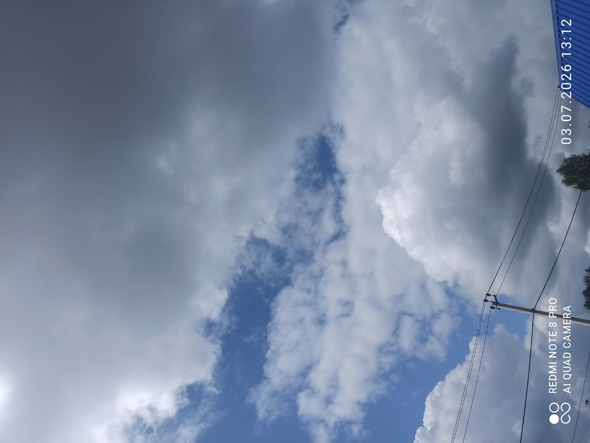 This is a landscape photograph taken outdoors in Sovetskiy, Russia, on July 3, 2026, at 13:12. The image is dominated by a sky filled with a mix of blue sky and dramatic clouds, ranging from bright white to dark gray. A utility pole with power lines is visible in the lower half of the frame, extending upwards towards the sky. The bottom edge of the image shows a glimpse of what appears to be a blue corrugated roof of a building and the tops of some trees. A watermark in the lower left corner reads "REDMI NOTE 8 PRO AI QUAD CAMERA," and the date and time stamp "03.07.2026 13:12" is located in the lower right corner. No people are visible in the image.
