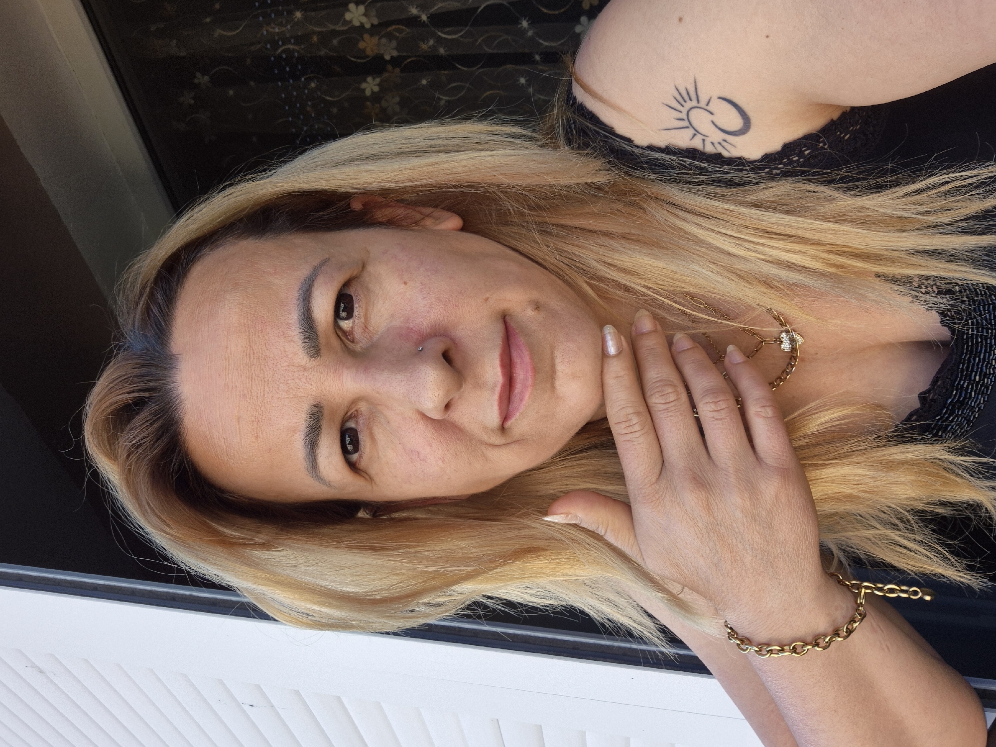 A medium shot, from the chest up, captures a Caucasian woman with blonde hair and light skin. She is looking directly at the camera. Her right hand is raised to her chin, with her fingers lightly touching her jawline. She is wearing a gold bracelet on her left wrist and a gold necklace with a heart-shaped pendant. On her right shoulder, a black tattoo of a crescent moon and sun is visible. Her clothing is a black top with lace trim. The background is split between a white paneled structure on the left and a dark curtain with a floral pattern on the right. The lighting suggests an outdoor setting during the day.