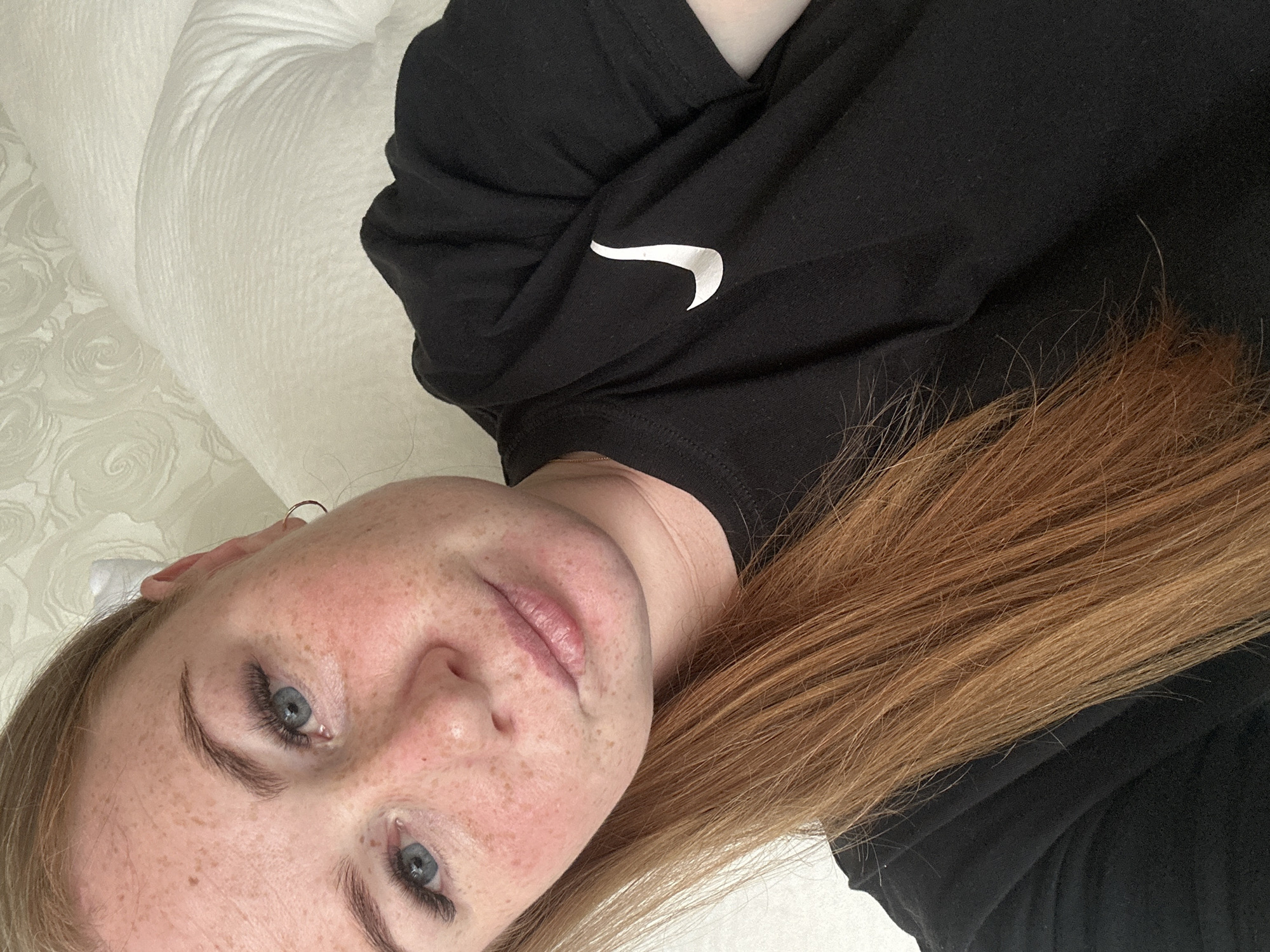 A young Caucasian woman with long auburn hair and fair skin dotted with freckles is lying down, looking towards the camera. She has blue eyes, full lips, and is wearing a black t-shirt with a white Nike logo on the chest. She has on long false eyelashes and a small hoop earring in her left ear. The background consists of a textured white wall with a subtle floral pattern and a white pillow. There is no identifiable location within the city of Chaikovsky, Russia.