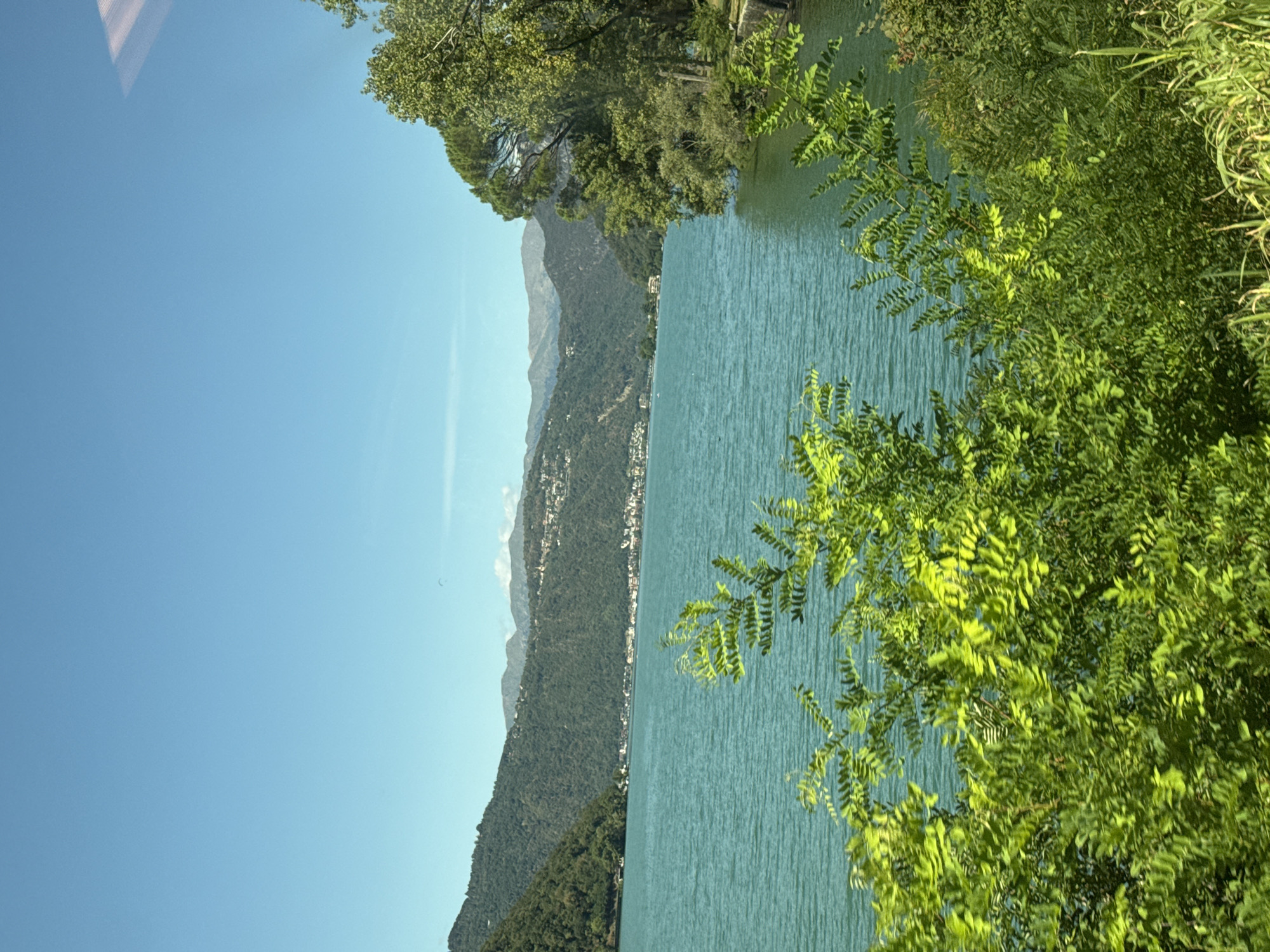 This is a landscape photograph taken from a slightly elevated vantage point, looking across a body of water towards a distant shore and mountains. The foreground is dominated by vibrant green foliage, obscuring the lower part of the scene and creating a natural frame. The water, likely a lake given the mountainous backdrop, is a striking turquoise blue and appears calm with gentle ripples.
In the mid-ground, a settlement is visible nestled at the base of lush green hills that rise into more rugged, hazy mountains in the distance. The architecture of the settlement appears to be of European style, with white and light-colored buildings. The sky is a clear, bright blue, suggesting a sunny day. The overall impression is one of natural beauty and tranquility. The location is identified as being in the city of Val Mara, Switzerland.