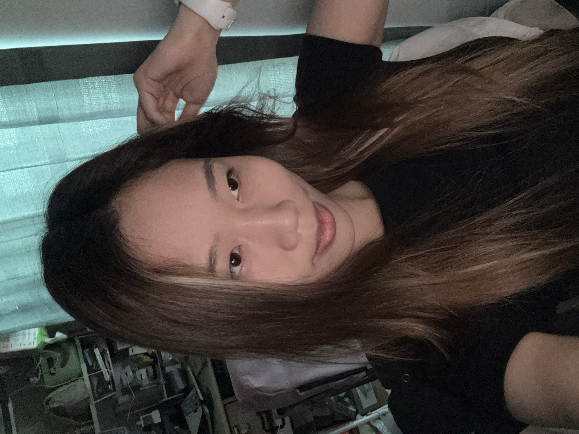 The image shows a young Asian woman with long, dark brown hair with blonde highlights, wearing a black t-shirt. She is seated in what appears to be a room with shelves of items in the background and a teal curtain. Her right hand is raised to her head, and she is wearing a white smartwatch on her left wrist. The scene suggests a casual indoor setting, possibly a home office or study area, located in the city of Nan'an District, China.