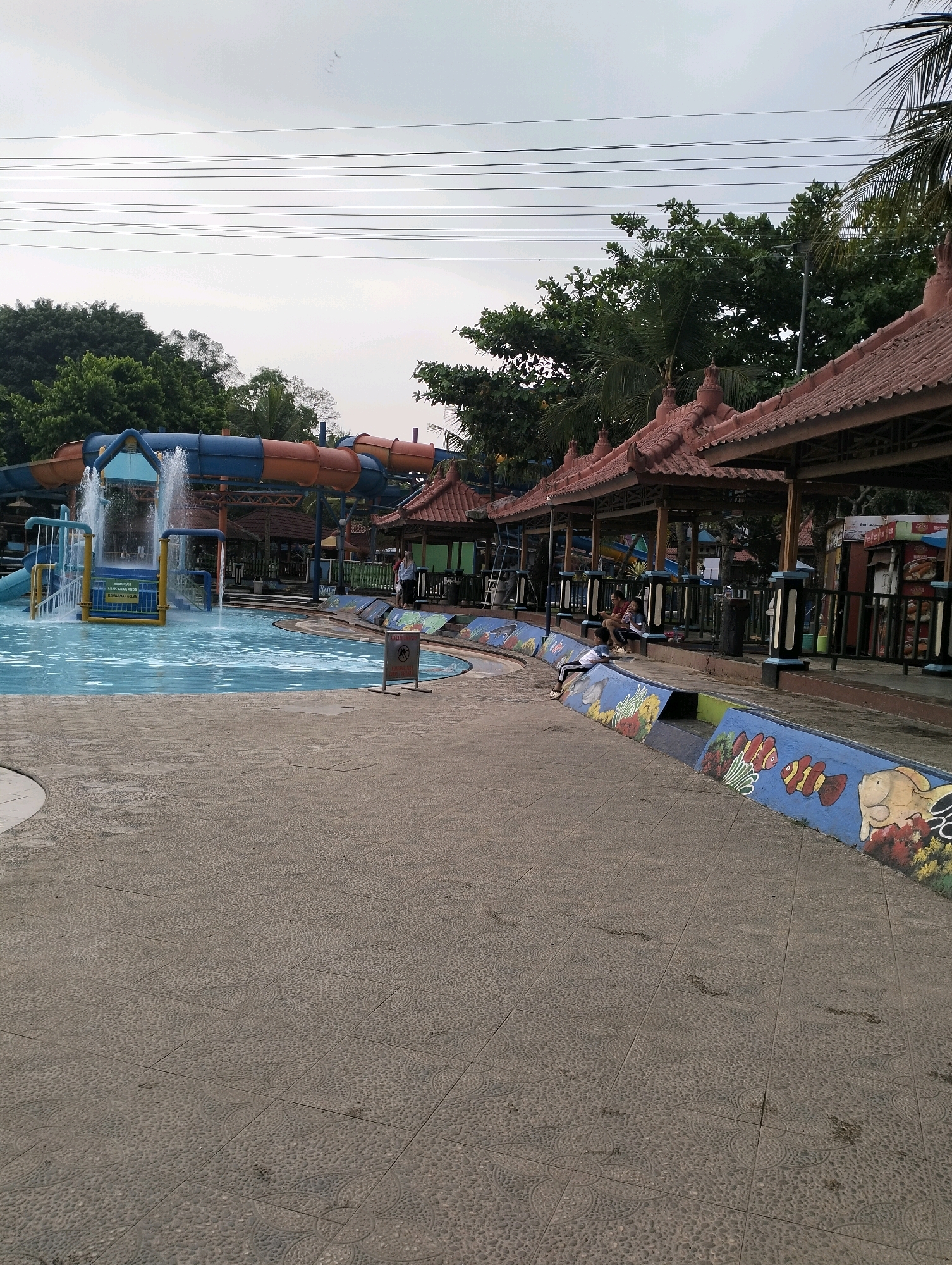 The image depicts an outdoor water park in Karangbanjar, Indonesia. In the background, a large water slide complex with blue and orange tubes is visible, with water spraying from a structure in the pool. To the right of the pool area, there are several open-air gazebos with traditional tiled roofs, providing shade. A few people are seated in these gazebos and along the pool's edge. The ground in the foreground is paved with textured tiles. Along the edge of the pool, a colorful mural of underwater life, including fish and coral, is painted on a low wall. The sky is overcast.