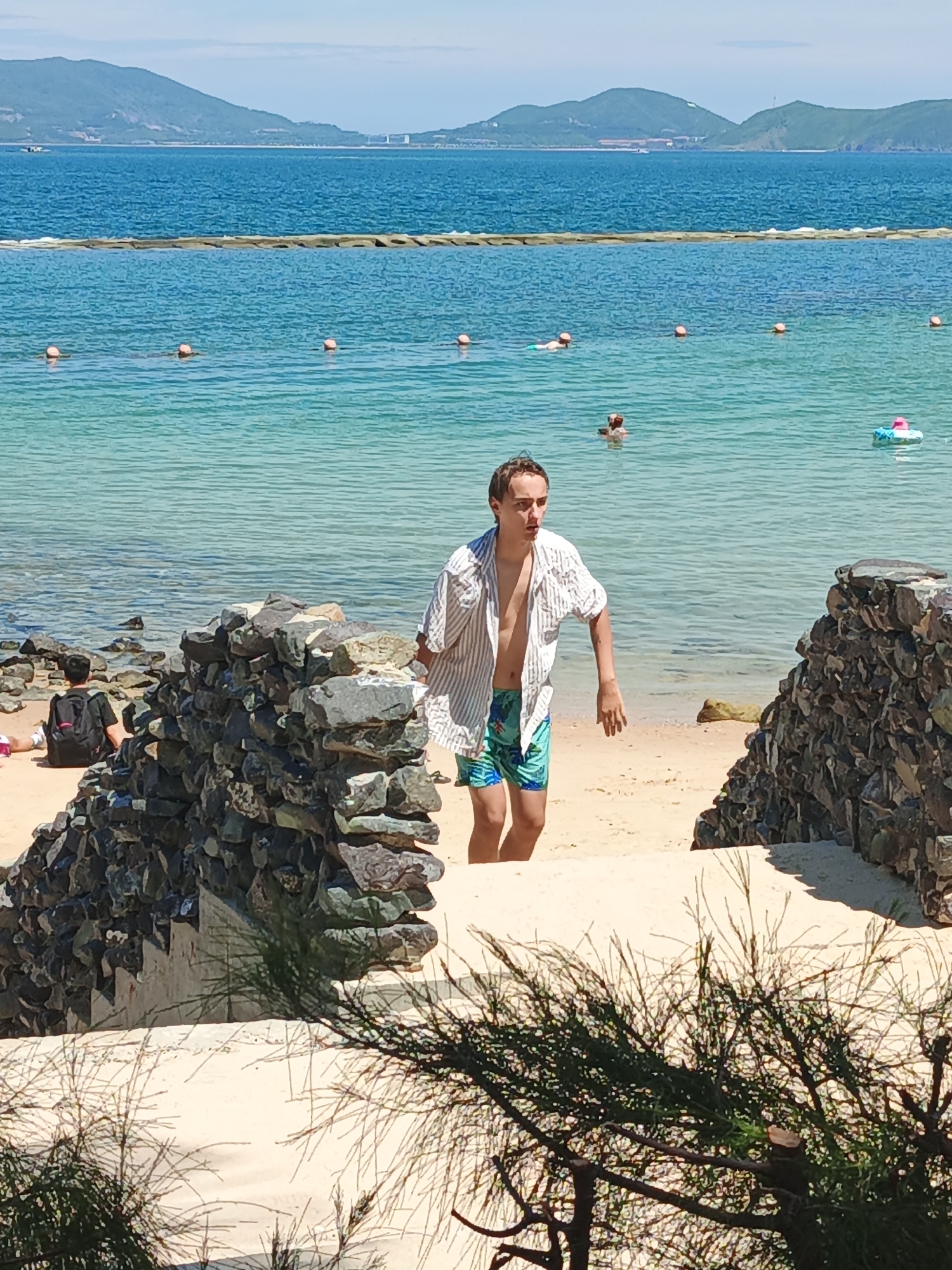 A young man in a striped open shirt and patterned swim trunks walks down a sandy path towards a beach. He is looking to his left with a surprised expression. In the background, the ocean is visible with several people swimming and a few buoys. A rocky wall is on the right side of the image, and some vegetation is in the foreground.