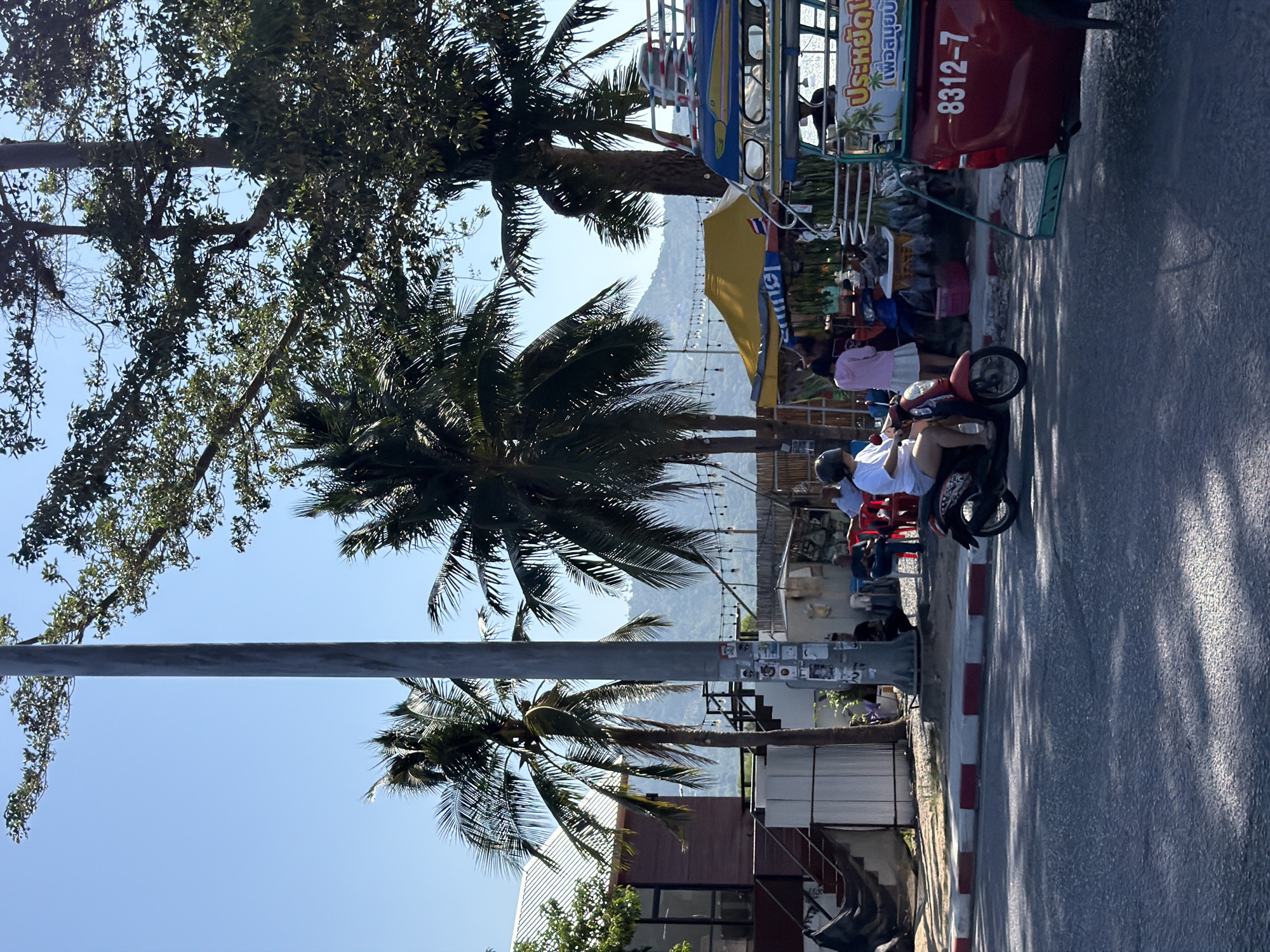 In the city of Ko Samui District, Thailand, a scene unfolds on a sun-drenched street lined with palm trees and a backdrop of hazy mountains. The foreground shows a paved road with distinct red and white curb markings, shaded by dappled sunlight filtering through the leaves. A red tuk-tuk, a three-wheeled vehicle common in Thailand, is parked on the right side of the frame, its license plate reading "8312-7" and adorned with Thai script.
To the left of the tuk-tuk, a person wearing a white shirt and shorts rides a red scooter, navigating the road. They are wearing a helmet. Further down the street, a yellow awning covers a small market stall, where a woman in a pink hoodie and a white skirt stands. In the background, a building with a white roof and brown siding is visible, featuring an outdoor staircase. Numerous string lights are suspended between poles, adding a festive touch to the scene. Several people are seated on chairs near the market, and more palm trees are scattered throughout the environment, contributing to the tropical ambiance. The sky is a clear, bright blue.