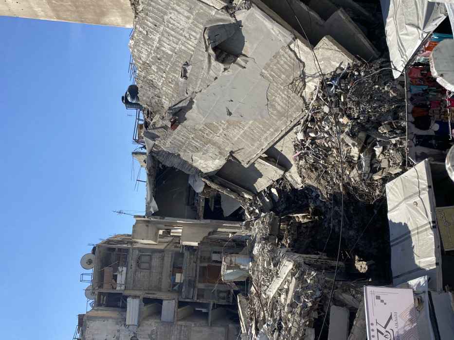 The image depicts a scene of extensive urban destruction in Khan Yunis, Palestinian Territory, under a clear blue sky. In the foreground, two market stalls with white canopies are partially visible; the left stall features a sign with Arabic text and "Ibra Alkhali," while the right stall displays various colorful clothing items. Immediately behind these stalls and filling the midground is a vast pile of pulverized concrete, twisted rebar, and general debris from collapsed multi-story buildings. Several large, gray concrete slabs are leaning at steep angles amidst the rubble, indicating severe structural failure. A deep, dark void is visible within the debris in the lower mid-section. To the left, a multi-story building, appearing structurally compromised but largely standing, features multiple windows, balconies, and visible interior sections. Its rooftop is equipped with satellite dishes and water tanks. To the right, on a partially collapsed section of a building, a black water tank is visible, and one individual in dark clothing is positioned near it. On the far right, a tall, plain concrete or masonry structure is partially visible. No overt actions or interactions between individuals are discernible, apart from the implied commercial activity of the market stalls adjacent to the destruction.