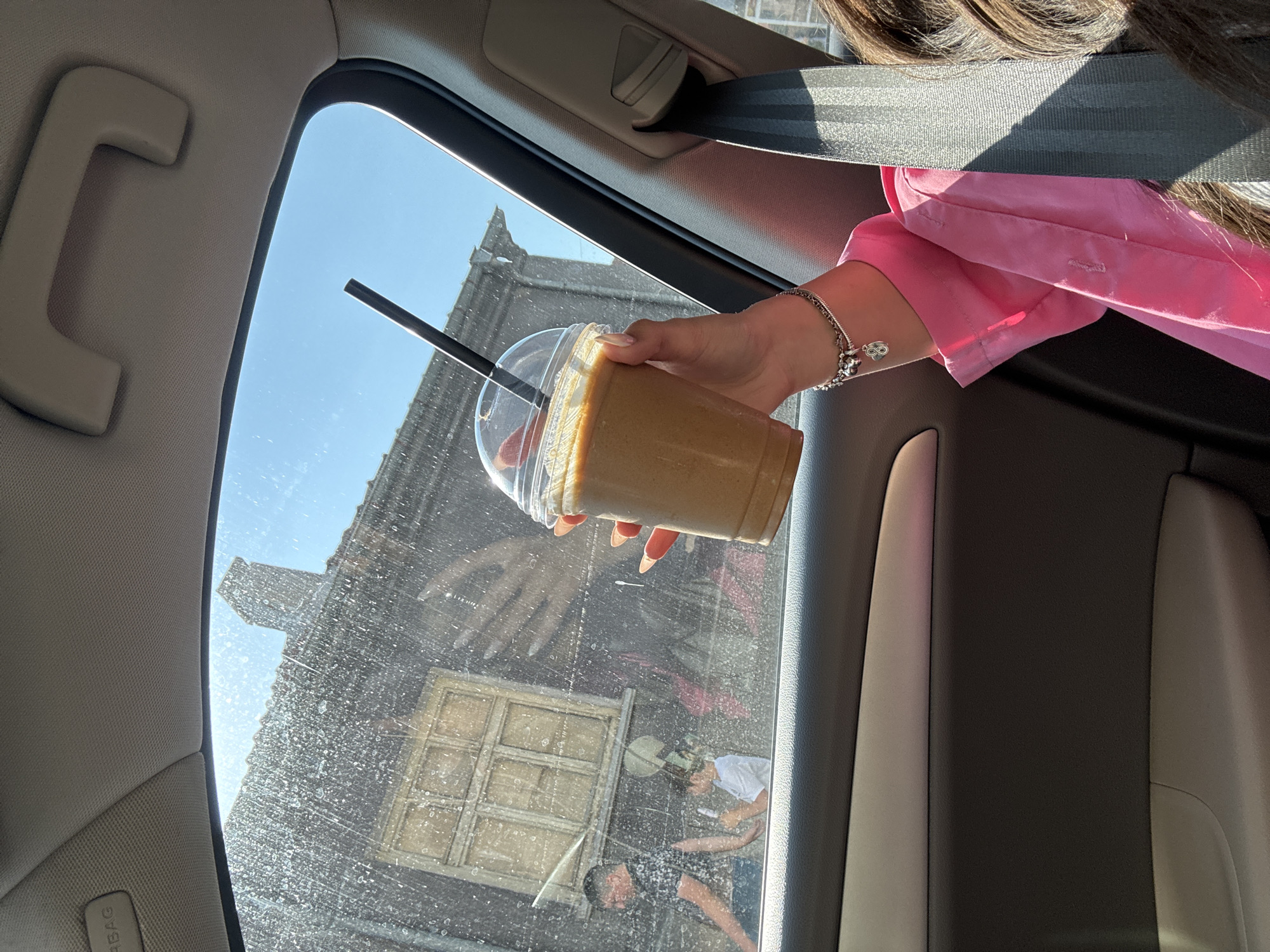 The image depicts a scene observed from the interior of a vehicle, looking out a side window towards a street in Rakovski, Bulgaria, on a brightly sunny day. The car window is significantly obscured by numerous water spots or dirt particles.
Inside the vehicle, a person's right arm, wearing a bright pink long-sleeved shirt, extends into the frame from the lower right. The wrist is adorned with a silver-colored charm bracelet featuring multiple charms, including an infinity symbol. The hand, with long, pointed nails painted with light orange/pink polish, firmly grasps a clear plastic cup containing a light brown/tan liquid and a black straw. Above the window, parts of the car's interior ceiling, including a light beige fabric headliner, a grab handle, and a visible "AIRBAG" label on the sun visor/pillar area, are visible. A dark grey seatbelt runs diagonally from the upper right to the lower right, partially obscuring the person's torso. Dark brown hair is visible in the lower right corner, indicating the presence of a passenger. The interior door panel, featuring beige and dark grey sections, is also visible below the window line.
Outside the car, through the speckled window, an older, weathered building with dark siding is prominent. It features a single, prominent wooden window frame with multiple panes, centered horizontally in the view. The building has a dark, tiled roof and a visible chimney against a clear, bright blue sky in the upper right.
Two individuals are visible outside the building. To the left, a man wearing a dark, patterned short-sleeved shirt and dark trousers walks from left to right. To his right and slightly behind him, a child, possibly a boy, stands wearing a white T-shirt and what appears to be a green or patterned hat/headband with earflaps. The child is holding a white object, possibly a mobile phone or toy, and appears to be looking down at it. The overall environment outside suggests an older urban or semi-urban setting under direct sunlight.