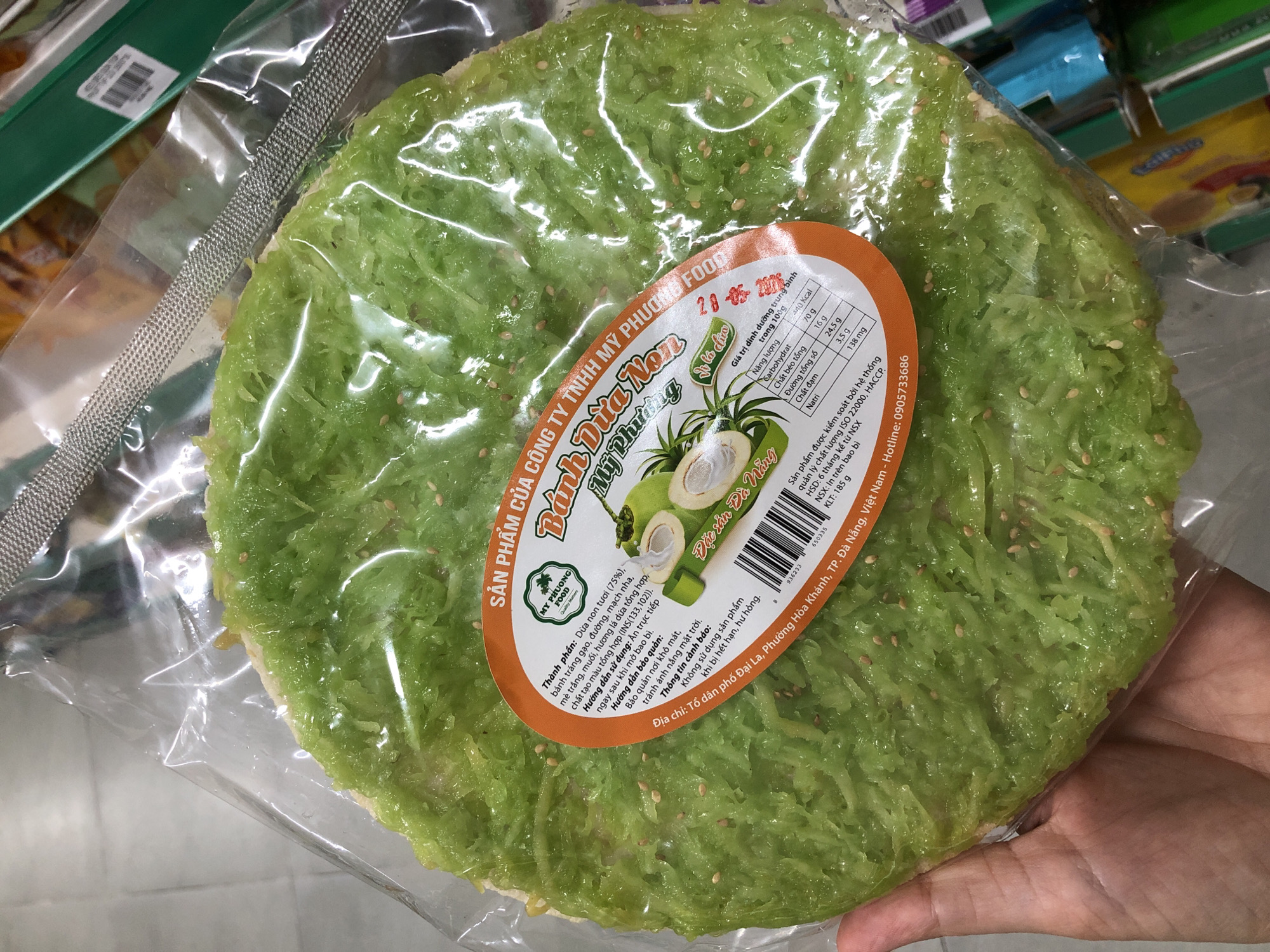 This is a close-up, eye-level media file of a person's hand holding a packaged food item. The food item is a round, flat, green confection, possibly a type of crispy pancake or cracker, made from shredded coconut and other ingredients. It is sealed in a clear plastic wrapper, allowing the texture and color of the food to be clearly visible. Scattered sesame seeds are visible throughout the green mixture.
A prominent orange and white circular label is affixed to the center of the packaged food. This label contains various text elements in Vietnamese, indicating product details and nutritional information. Visible text includes "SẢN PHẨM CỦA CÔNG TY TNHH MỸ PHƯƠNG FOOD," "MỸ PHƯƠNG FOOD," "Đặc sản Đà Nẵng," and a nutritional breakdown per 100g, listing calories, carbohydrates, fat, sugar, protein, and sodium. It also mentions manufacturing and expiration dates, quality control systems (ISO 22000, HACCP), and a contact hotline. The "Địa chỉ" (address) section specifies "Tổ dân phố Đại La, Phường Hòa Khánh, TP. Đà Nẵng, Việt Nam."
The background is out of focus but suggests a retail environment, possibly a grocery store or market, with shelves and other products visible. The lighting appears to be artificial, typical of an indoor setting. The person's hand is visible at the bottom right, gently supporting the packaged item. The overall impression is a product shot intended to showcase a specific Vietnamese food item.