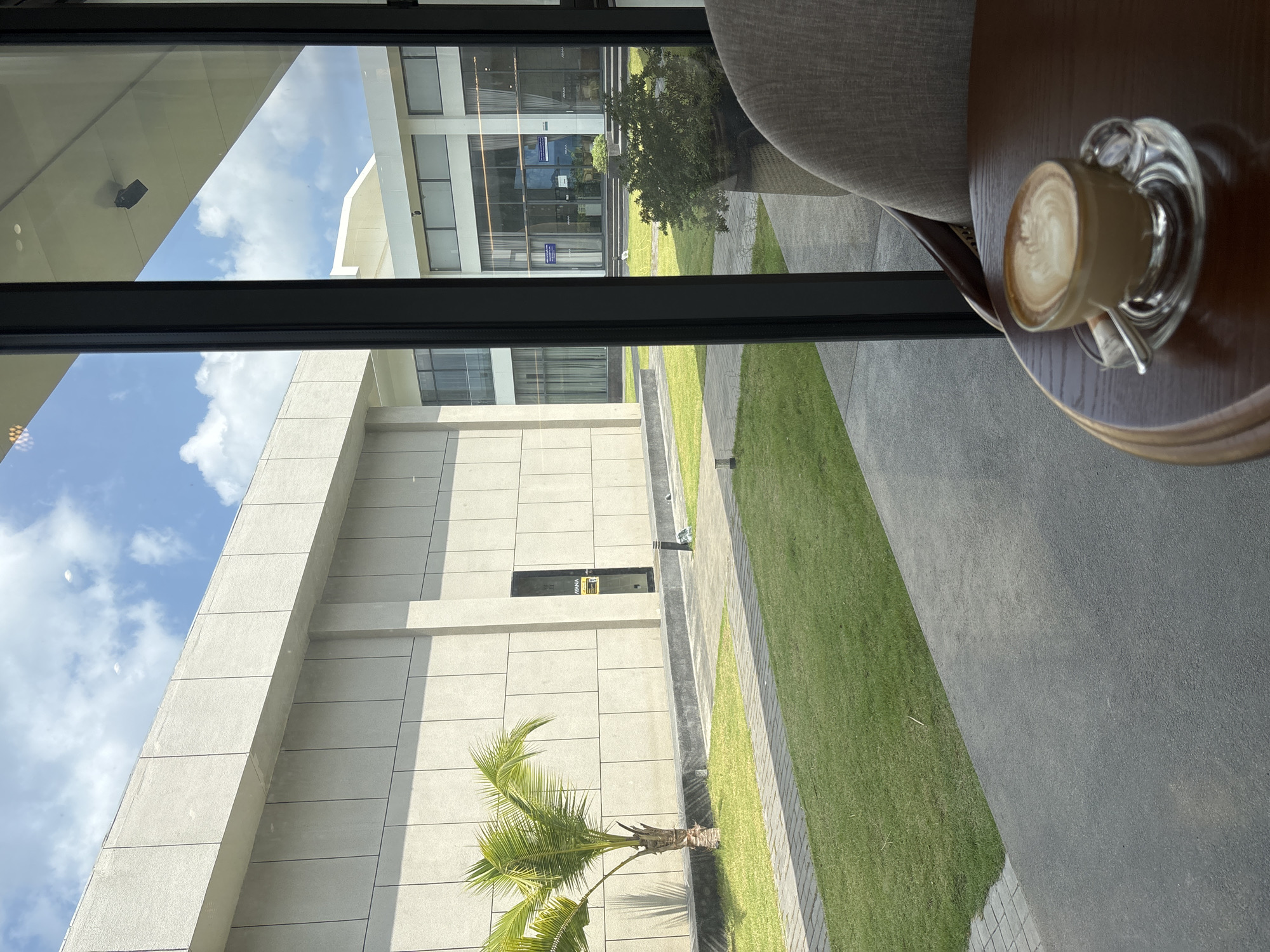 A view from inside a building in Choeng Thale, Thailand. The scene is seen through a window, with a cappuccino in a glass cup on a round wooden table in the foreground. A grey chair is partially visible in the top right corner. Outside the window is a modern building with beige tiled walls, a small palm tree, and patches of green lawn interspersed with paved walkways. Further away, another building with glass windows and doors is visible. The sky is blue with scattered clouds.