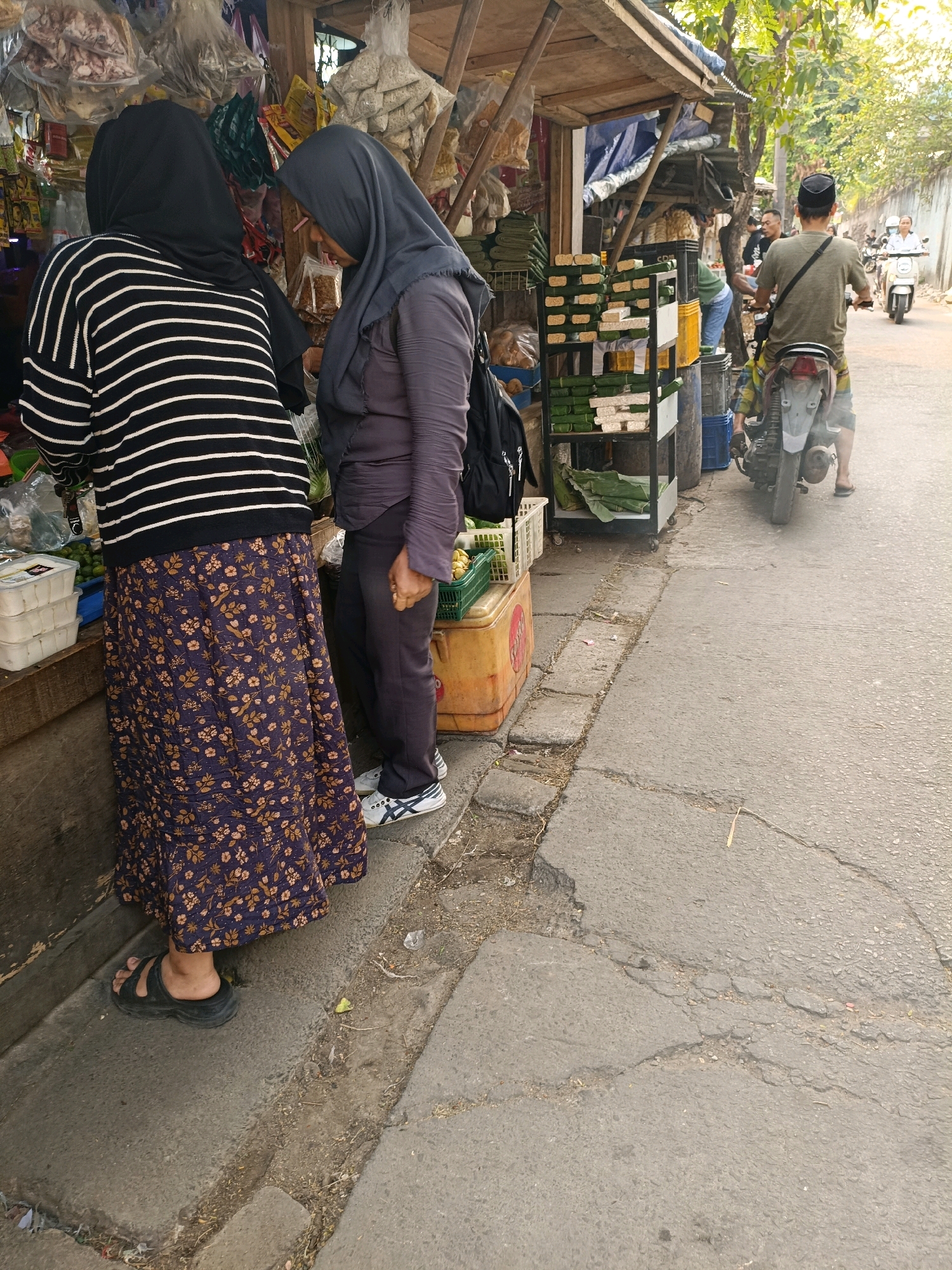 The image depicts a street scene in Tangerang, Indonesia, focusing on a roadside stall and its surroundings. Two women, dressed in hijabs and modest attire, are standing near the stall, appearing to be engaged in a transaction or discussion. The woman on the left wears a black and white striped sweater and a floral-patterned skirt, with black sandals. The woman on the right is in a dark purple hijab and matching pants, with white athletic shoes.
The stall itself is constructed from wood and covered with a blue tarp. It displays various goods, including packaged food items, possibly dried goods or snacks, some wrapped in green leaves, and plastic containers holding produce. Shelving units are visible, holding neatly arranged stacks of items. A yellow cooler box is also present.
In the background, a man wearing a green t-shirt and a black cap is riding a motorcycle away from the camera. Another person, also on a motorcycle, is visible further down the street. The road is paved and appears to be in moderate condition with some cracks and debris. Trees line the side of the road, casting shadows. The overall atmosphere suggests a typical daytime market or street-side vendor scene in an urban Indonesian setting.