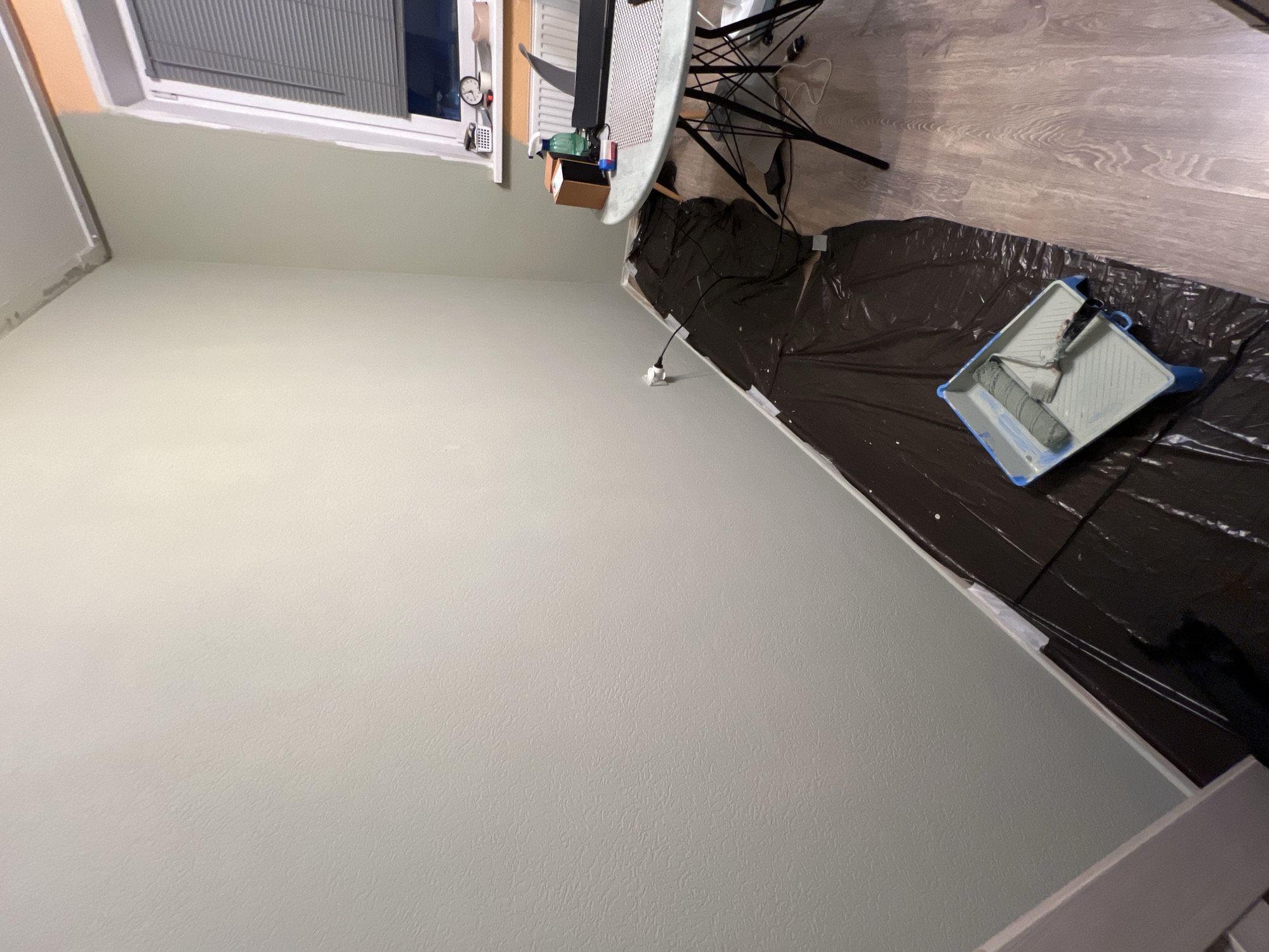 An interior room in Saint Petersburg, Russia, is depicted during or immediately after a painting project. The walls are painted a light sage green, and the ceiling is white. The floor is covered with light-colored wooden laminate, a significant portion of which is protected by black plastic sheeting taped along the white baseboards. In the foreground, on the plastic sheeting, a blue and white paint tray holds a used paint roller and a paintbrush. A white electrical outlet on the left wall has a black cord plugged in, which extends across the protected floor. To the right, a round table with a light grey top and black metal legs is positioned. On the table, a dark-colored laptop, a green spray bottle, a small brown box, and a red object are visible. A white patterned placemat is under these items. Above the table, a white window with closed grey Venetian blinds reveals a dark exterior. A white radiator is present below the window, and on the windowsill, various items including a white organizer, a clock, and a roll of white tape are arranged. No individuals are present in the scene.