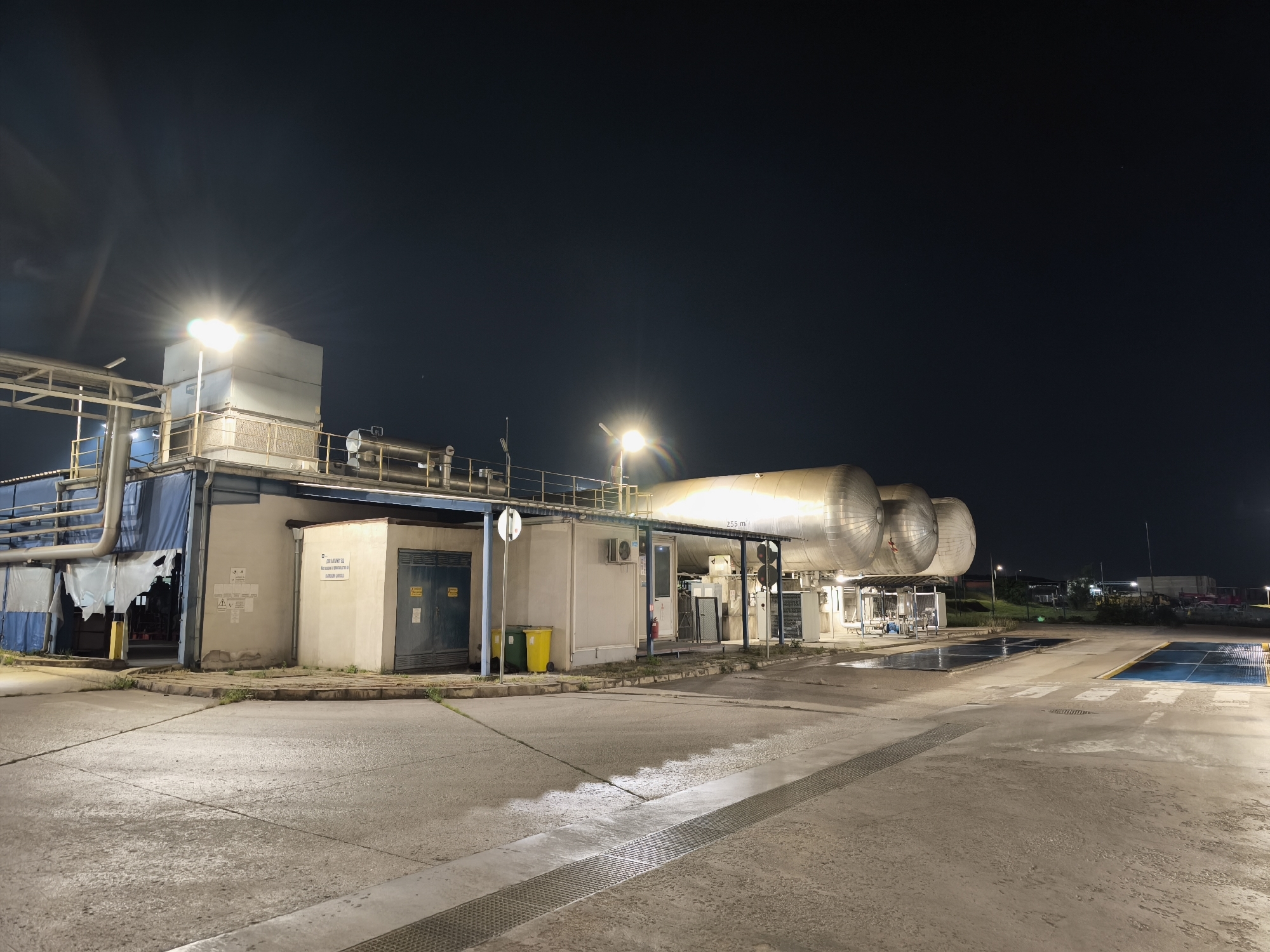 The image depicts an industrial site at night. Several large, horizontal cylindrical tanks dominate the scene's right side, illuminated by overhead lights. To the left of the tanks, a building with multiple sections is visible. The building appears to be a combination of enclosed rooms and open, covered areas with pipes and equipment on the roof. A yellow trash can stands near one of the building sections. The ground is paved and wet in places, reflecting the artificial light. The sky is dark, suggesting it is nighttime. No people are visible in the scene. The location is Unknown, Unknown.