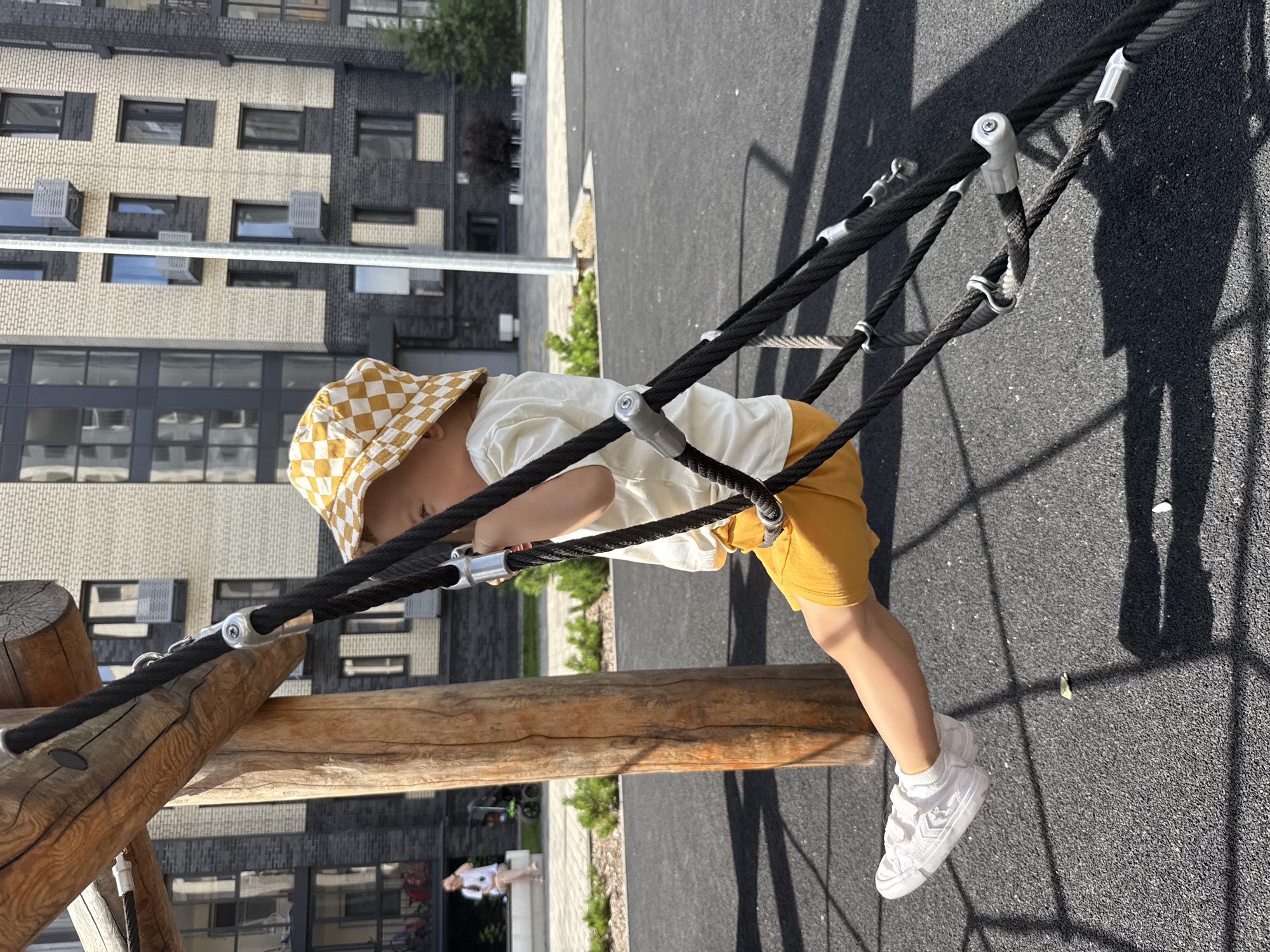 A young child wearing a yellow and white checkered bucket hat, a light-colored t-shirt, and yellow shorts is playing on a playground structure. The child is positioned on a climbing net made of thick black ropes and is holding onto the ropes. The playground is situated on a dark gray rubberized surface, with shadows from the climbing structure cast across it. In the background, a modern apartment building with a light beige and dark gray brick facade is visible. To the left of the building, a few trees and a woman in a white shirt and shorts can be seen. The overall scene depicts an outdoor recreational area within an urban or suburban setting, possibly in Rusanovo, Russia, given the context.