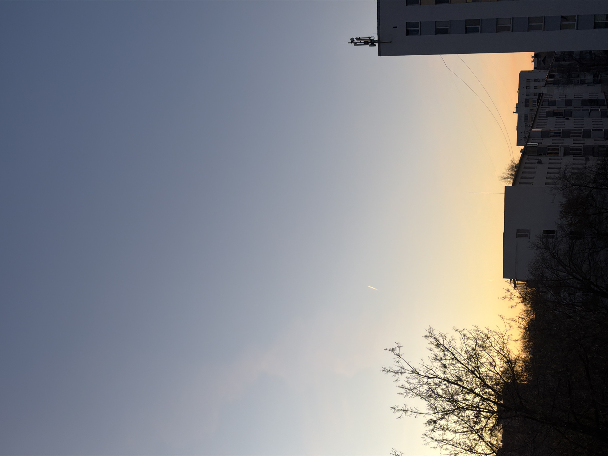 The image shows a view of the sky at sunset in Naberezhnye Chelny, Russia. The sky transitions from blue to a warm yellow-orange near the horizon. Several apartment buildings are visible on the upper right and along the bottom edge of the image. A communication tower is affixed to the roof of one of the buildings. Bare trees are silhouetted against the sky along the lower right. A faint contrail from an airplane is visible in the center of the frame. Power lines are also visible.