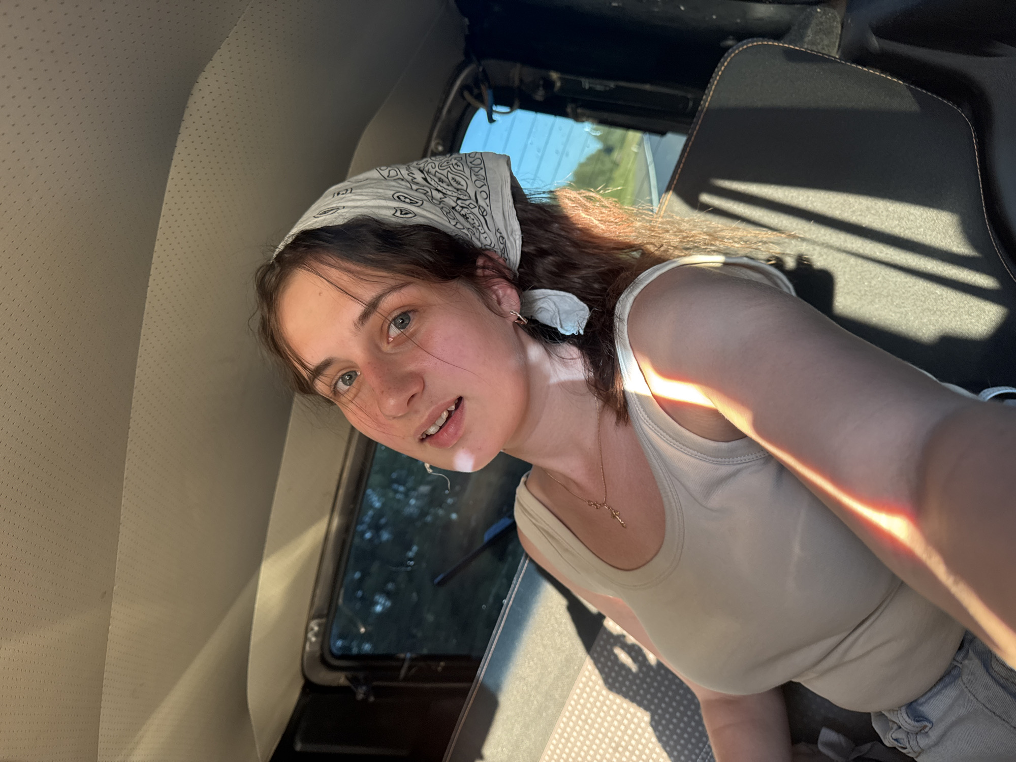 The image shows a young woman sitting in the back seat of a vehicle, with the sun illuminating her face and chest. She is wearing a white bandana on her head, a light-colored tank top, and appears to be wearing denim shorts. A necklace with a small cross pendant is visible around her neck. Her hair is dark brown and wavy. The interior of the vehicle is visible, with textured headliner and dark-colored seats. Through the windows, trees and a bright blue sky can be seen, suggesting an outdoor setting, possibly on a road trip. The lighting and shadows indicate it is daytime. The location in Bol'shaya Sosnova, Russia, is not discernible from the image itself.