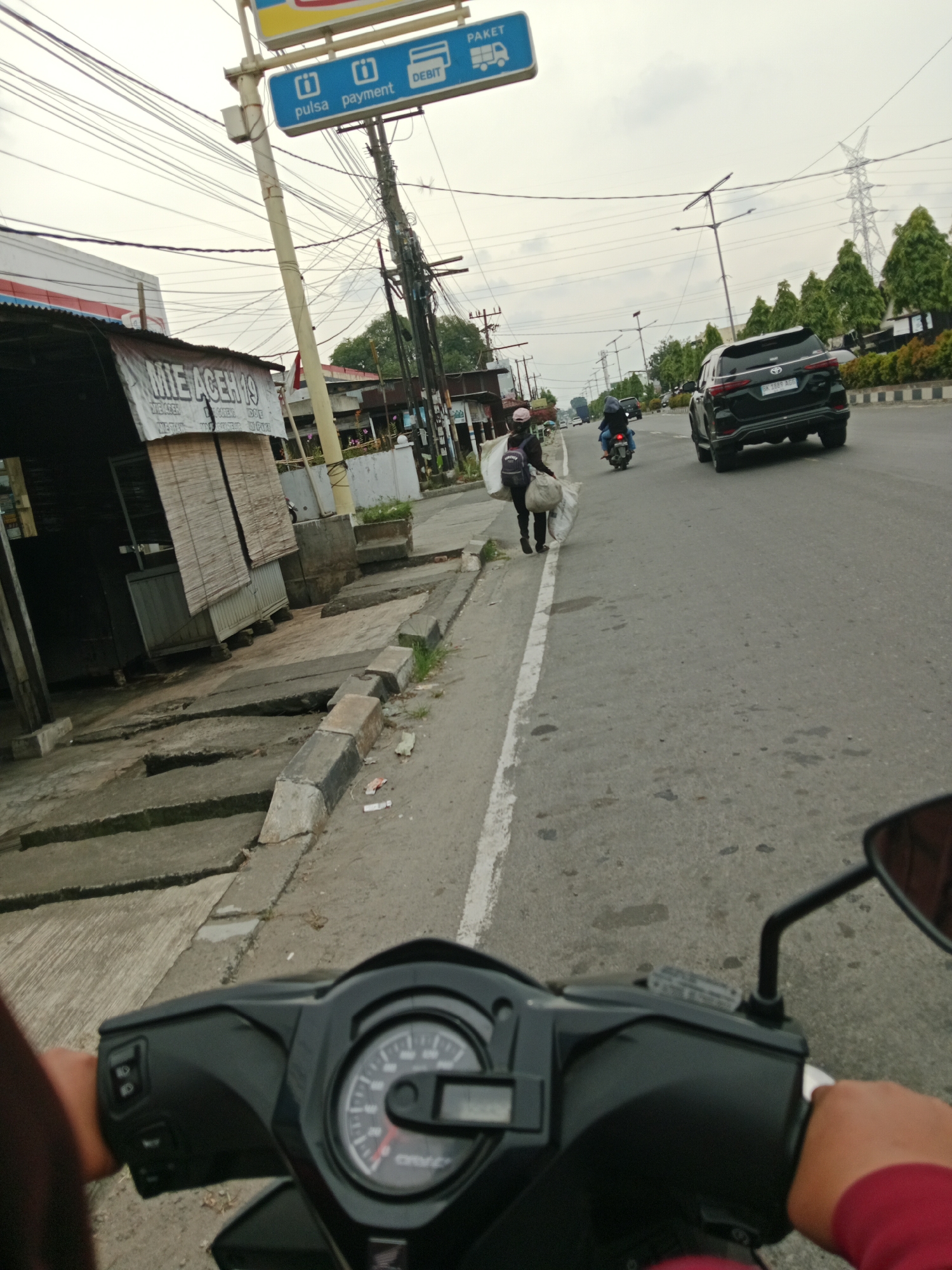 The image captures a first-person perspective from a motorcycle on a street in the City of Binjai, Indonesia. The foreground shows the handlebar and dashboard of the motorcycle, with a speedometer indicating a moderate speed. In the middle ground, a black SUV with license plate "BK 1869 ADF" is traveling on the road. To the left, a person walks along the sidewalk carrying two large sacks. Further down the road, another person is seen riding a motorcycle. Overhead, a complex network of electrical wires crisscrosses the sky. The environment appears to be urban with buildings and trees visible along the side of the road. A signpost with "PAKET" and "DEBIT" markings is also present. The sky is overcast, suggesting a cloudy day.