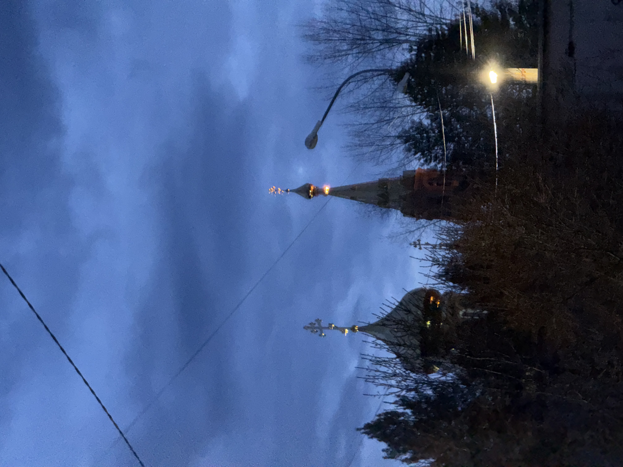 The image depicts a nighttime scene with a dark, cloudy sky. Two illuminated spires of a building or church are visible, one slightly obscured by trees. A street lamp and electrical wires are also present. The location is identified as being in the city of Unknown, Unknown.