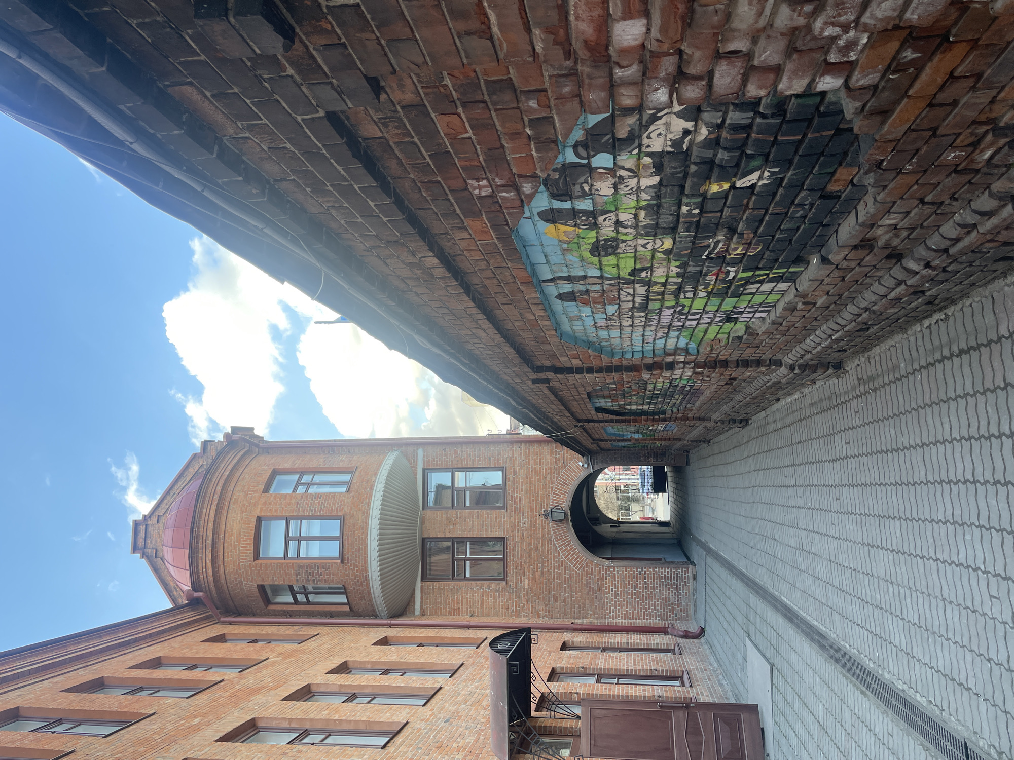 The image depicts a narrow alleyway in Krasnoyarsk, Russia. The alley is paved with grey interlocking bricks and is flanked by tall brick buildings. The buildings are primarily constructed of red brick, with a mix of architectural styles, including arched doorways, rounded balconies, and varying window styles. One building features a mural on the underside of the alley's roof, composed of smaller brick-sized tiles, depicting a group of figures. The sky is visible at the end of the alley, with blue skies and scattered clouds. The perspective of the image is looking up the alleyway towards the sky, emphasizing the height of the surrounding buildings.