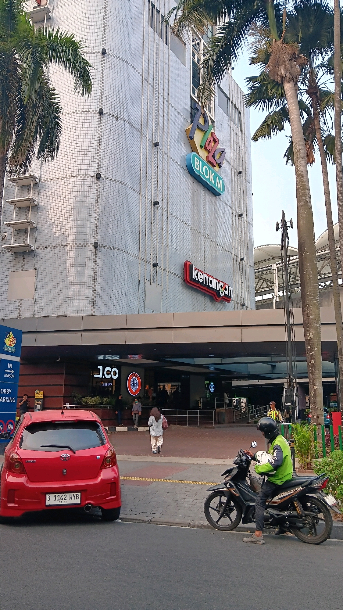 The image depicts the exterior of the Plaza Blok M shopping mall, located in the Kebayoran Baru district of South Jakarta, Indonesia. The building's facade is covered in light-colored mosaic tiles, with large windows visible at the upper levels. A prominent sign displaying "Plaza BLOK M" in colorful lettering is affixed to the building. To the right of this sign, another red neon sign reads "kenangan".
In the foreground, a red Toyota Yaris is parked on the street, facing away from the camera. Its license plate reads "B 1142 HYB". Adjacent to the car, a person wearing a green vest and a black helmet is seated on a black motorcycle, holding their helmet. The motorcycle is parked on the sidewalk.
Further back, pedestrians are seen walking into the mall entrance, which features prominent signage for "J.CO" and what appears to be a Starbucks. The area is paved with concrete and there are palm trees and other greenery lining the street and in front of the building. The sky is visible in the upper right portion of the image, appearing clear and bright, suggesting daytime. The overall scene captures a typical urban street view in front of a commercial establishment in Jakarta.