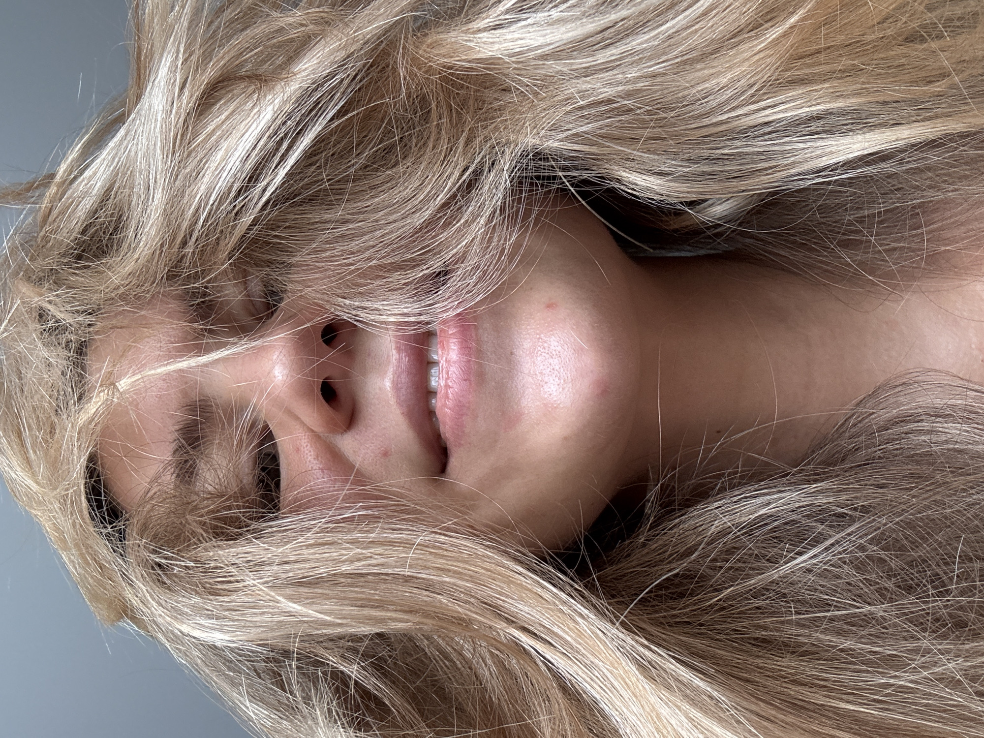 A close-up, slightly angled portrait shows a person with long, blonde hair partially obscuring their face. Their eyes are mostly hidden by the hair, but a portion of their nose, mouth, and jawline are visible. The background appears to be a neutral gray or white. The specific location and city are unknown.