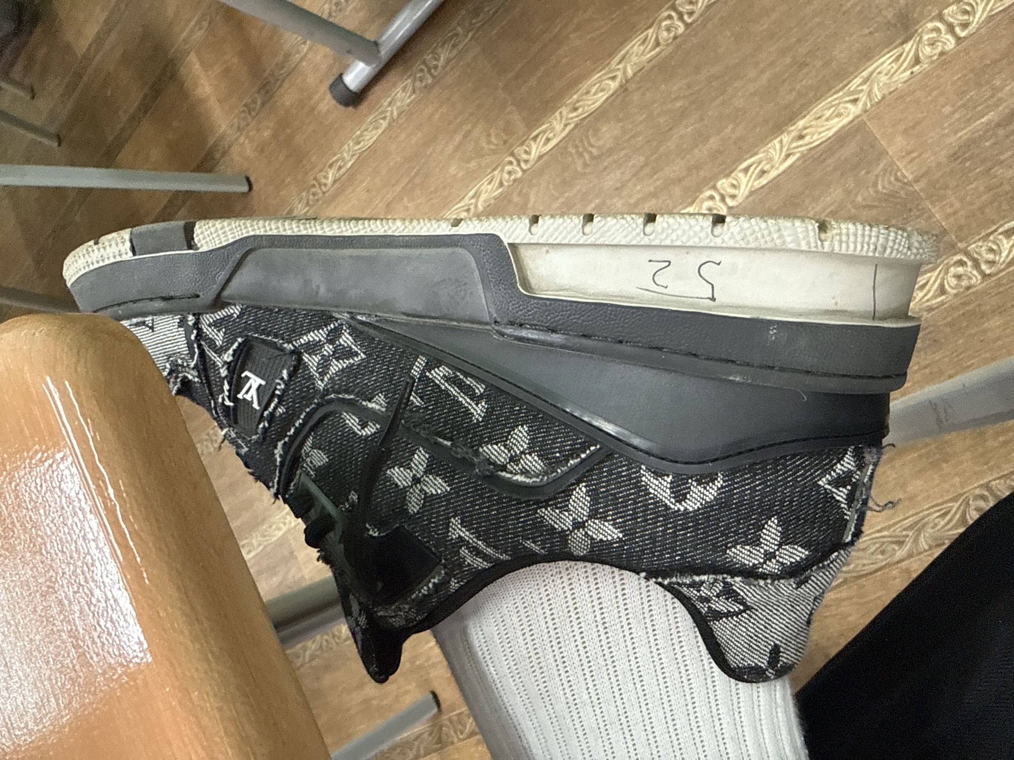 The image displays a close-up, angled view of a person's foot and lower leg, featuring a distinctive shoe and a white sock, in an indoor environment. The prominent subject is a sneaker-style shoe crafted from a dark, denim-like fabric patterned with a repeating design resembling the Louis Vuitton monogram, including stylized "LV" initials and four-petal floral motifs in white or light grey. The shoe possesses a multi-layered sole; the mid-sole is white, and a handwritten black number "52" is visible on its outer side. A white, ribbed sock covers the foot, and a portion of a bare leg, exhibiting a glossy sheen, is visible on the left.
The background features a light brown wooden or laminate floor, characterized by decorative patterns that suggest an inlaid design. Metal legs of what appears to be a chair or table are partially visible, indicating a typical indoor setting such as a classroom, office, or communal space. The lighting is soft and consistent with indoor artificial sources. The scene captures a casual moment, likely taken while the person is seated. This image was captured in Birobidzhan, Russia.