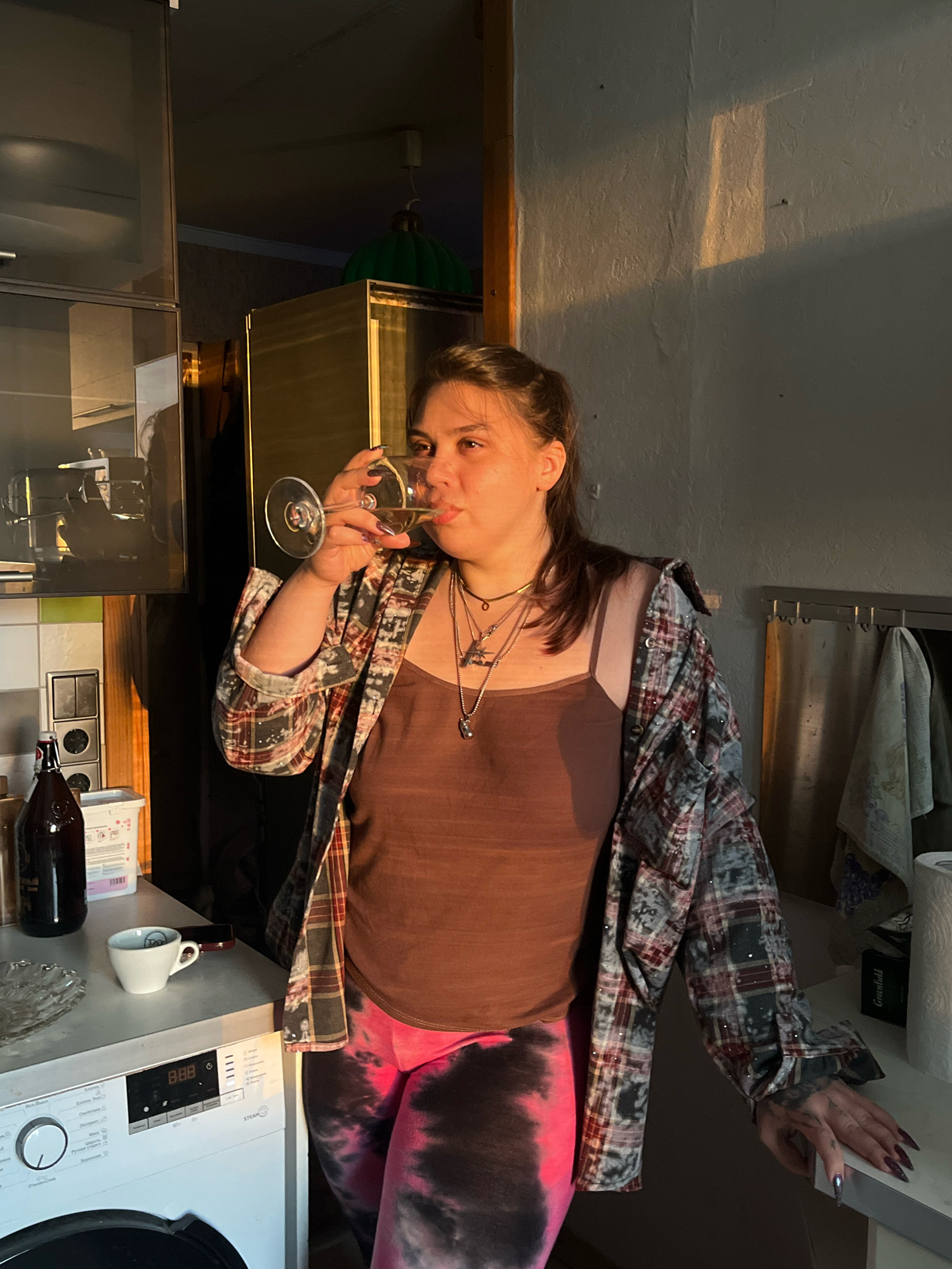 A medium shot captures a woman standing in what appears to be a kitchen, with a washing machine visible in the foreground to the left. She is holding a stemmed glass of white liquid to her lips and drinking from it. She is wearing a brown tank top, an open plaid shirt with reflective elements, and pink and black tie-dye leggings. Several necklaces adorn her neck. The lighting suggests it is late afternoon or early evening, with strong, warm sunlight casting shadows on the wall. Objects on the counter include a bottle, a small white cup, and a glass dish. The background shows kitchen cabinets and a green, dome-shaped object that could be a decorative item. The setting does not provide any specific visual cues to indicate it is located in Moscow, Russia.