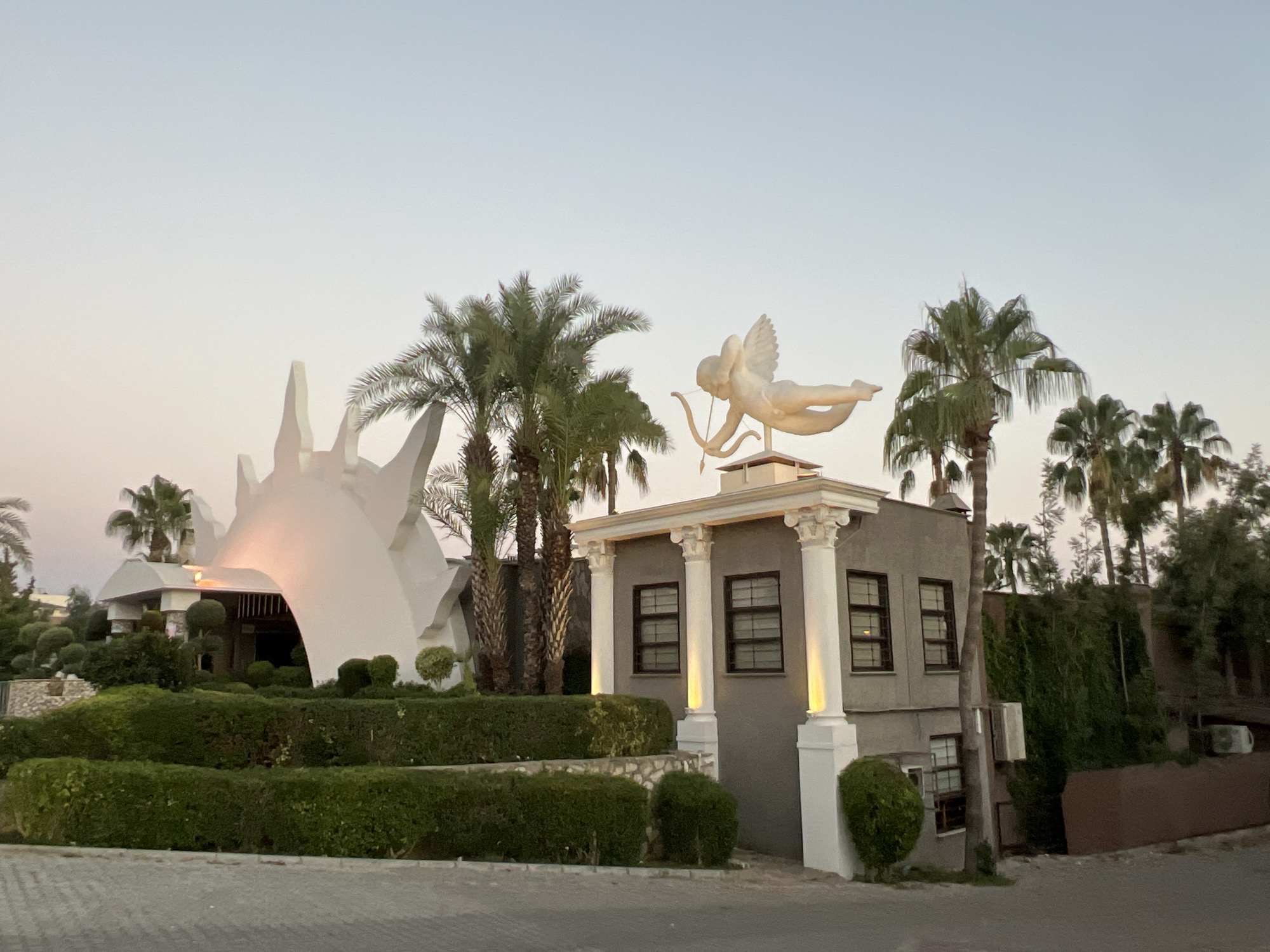 An exterior shot of a building in Alanya, Turkey, at dusk. The building features a white, abstract, sun-like sculpture on its left side and a statue of Cupid with a bow and arrow on its roof. The building is surrounded by palm trees and manicured hedges. The sky is a pale, gradient orange and blue, indicating the sun has recently set. The building's facade on the right is grey with white columns and dark-paned windows, illuminated by soft yellow lights.