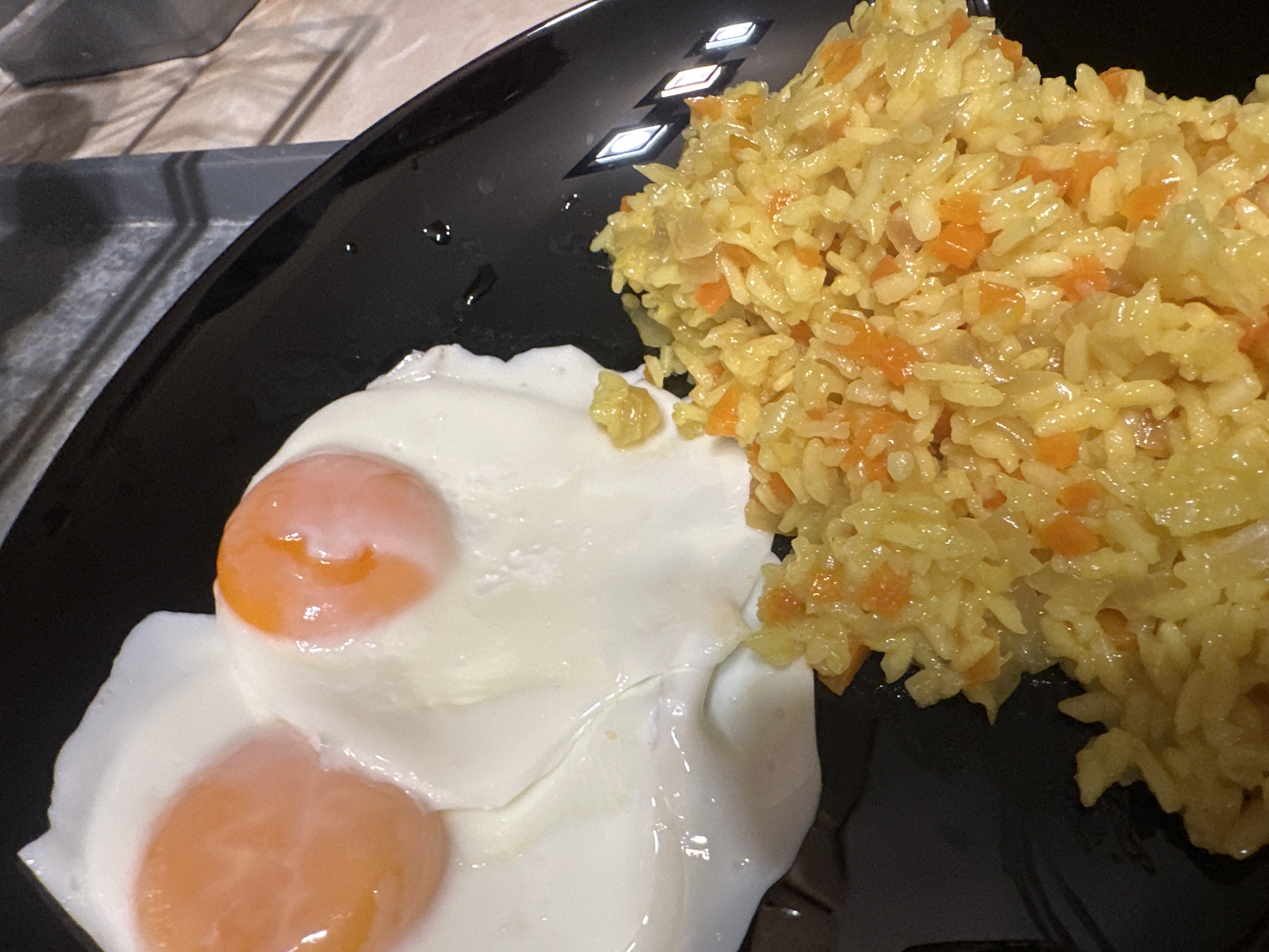 On a black plate, two fried eggs with bright orange yolks and opaque white albumen are positioned to the left of a pile of yellow rice pilaf containing small orange diced vegetables, possibly carrots. The plate's reflective surface shows overhead lighting, suggesting an indoor setting. The background includes a surface that could be a countertop or table with a subtle wood-grain pattern. The scene depicts a simple meal. There is no information to conclude this image was taken in Burgas, Bulgaria.
