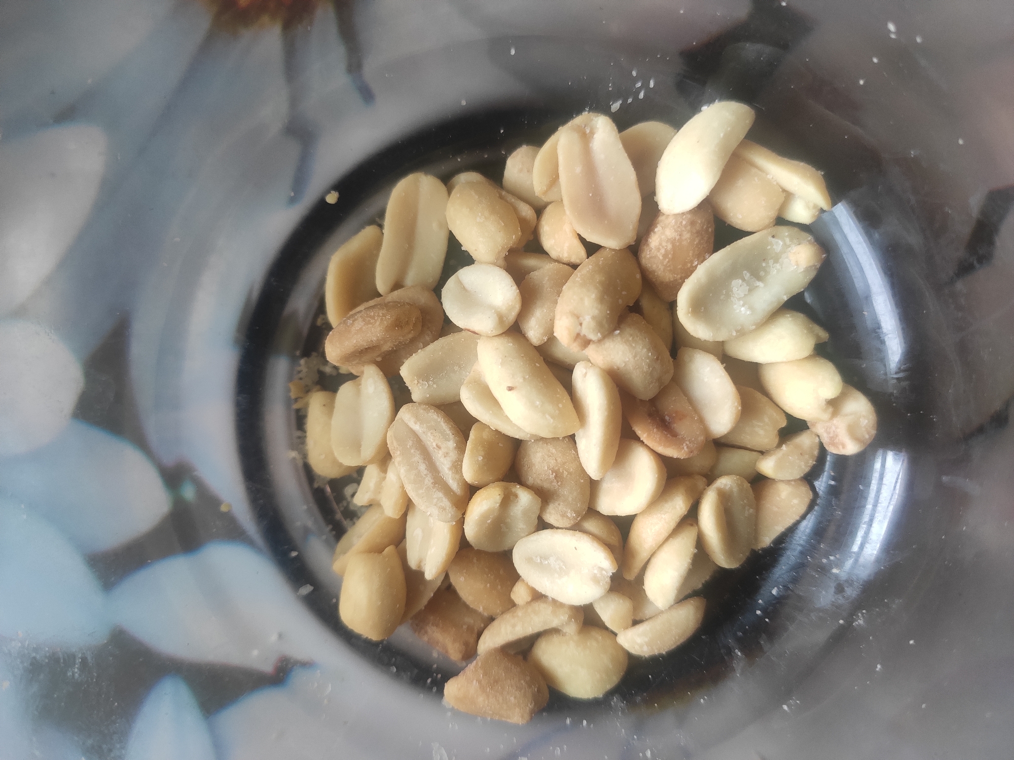 A close-up, top-down shot shows a pile of roasted peanuts inside a clear glass jar. The peanuts are light brown and have a slightly wrinkled texture. Some of them are broken in half, revealing their interior. Small white salt crystals are visible on some of the peanuts. The background inside the jar is blurred, showing a pattern of pale blue and white shapes that appear to be flowers. The exterior of the jar shows reflections of a floral pattern, possibly a tablecloth or wallpaper. The image does not contain any people or evidence of specific locations within the city of Kostopil, Ukraine.