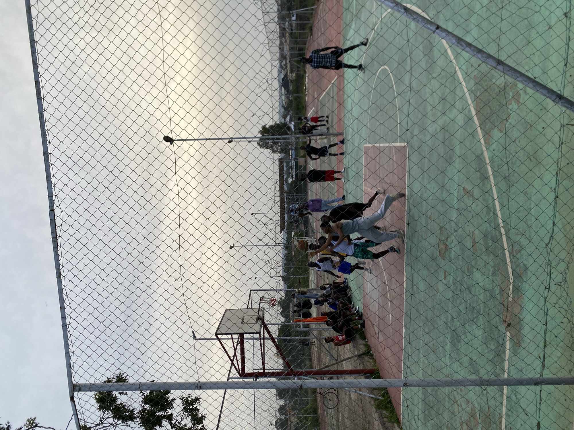 A group of young men is playing basketball on an outdoor court in Jos, Nigeria. The court is enclosed by a chain-link fence, and a basketball hoop with a backboard is visible. The sky is overcast, suggesting it may be late afternoon or early evening. Some players are actively engaged in the game, while others are observing from the sidelines or resting. The scene appears to be in a somewhat urban or semi-urban setting, with buildings and trees visible in the background. The overall atmosphere is casual and recreational.