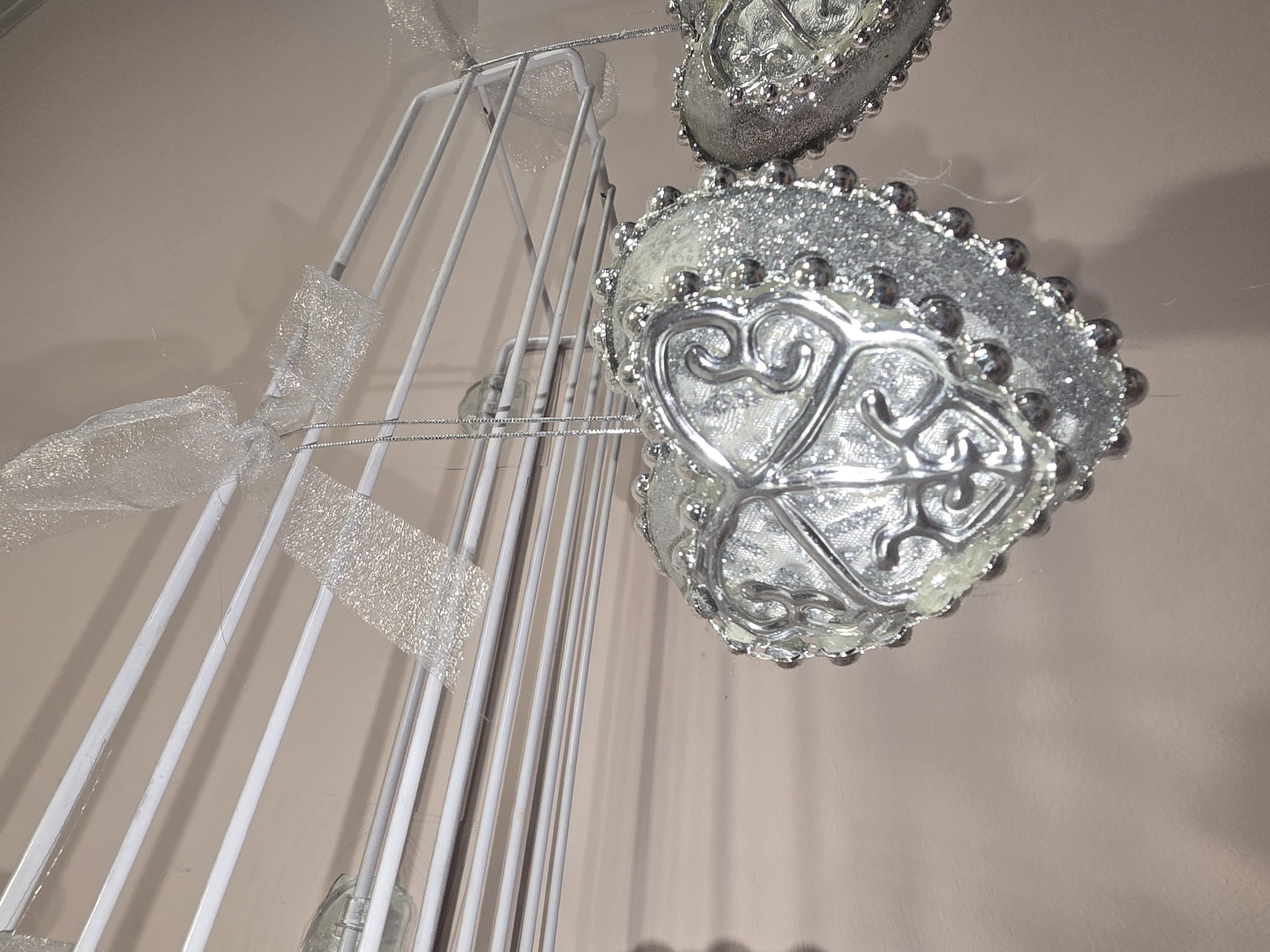 This is a close-up, eye-level shot of two silver, heart-shaped ornaments hanging from a white wire rack. The ornaments are decorated with glitter and small silver beads around their edges, and feature an ornate, swirling silver design in the center. A sheer, glittery ribbon is tied above the ornaments to the rack. The background is a plain, light pink wall. There are no people visible, and no identifiable landmarks suggest a specific location within Ulan-Ude, Russia.