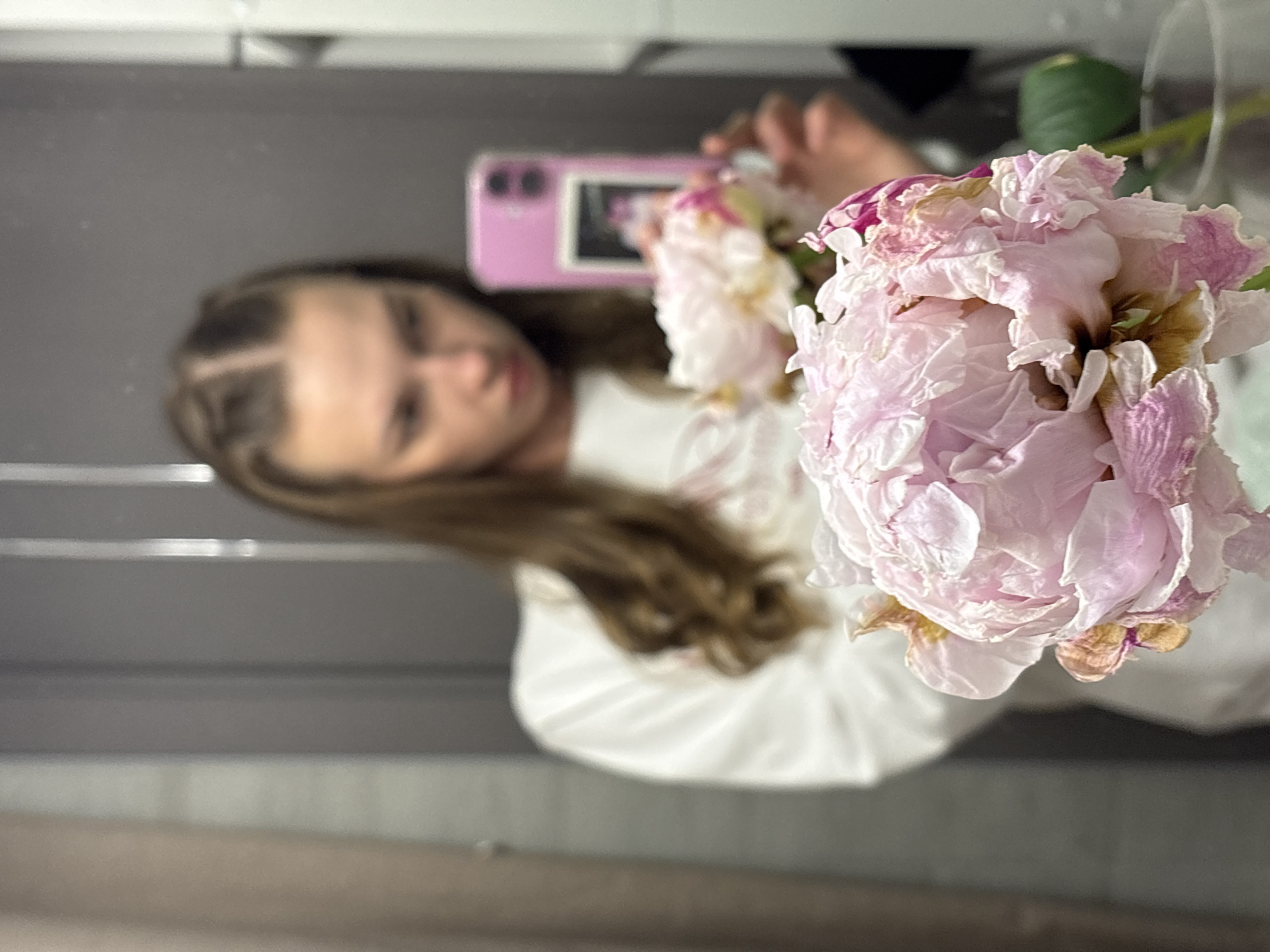 A young woman with brown hair, styled in two braids, is reflected in a mirror. She is wearing a white sweatshirt and holding a pink smartphone. In the foreground, two large pink peonies are visible, with one in sharp focus. The background appears to be a bathroom, with grey walls and a door. There are no visible indicators of the specific location within the city of Perm, Russia.