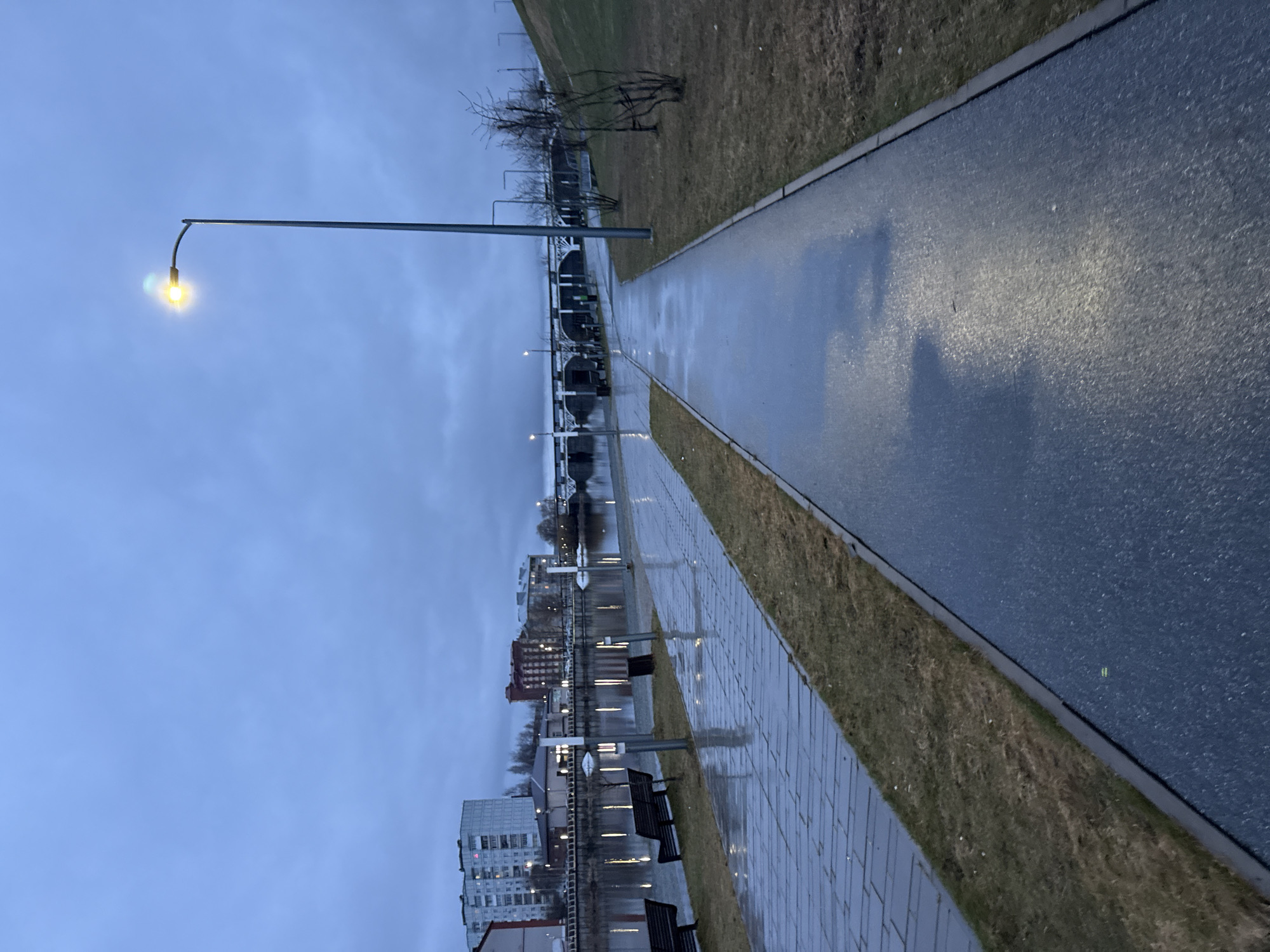 The image depicts a walkway beside a body of water, likely a river or lake, in Votkinsk, Russia. The walkway is made of two sections: a paved brick section and a smooth asphalt path. Both surfaces appear wet, indicating recent rain or damp conditions. Along the brick path are benches and light posts reflecting in the water. In the background, there are buildings, including what appears to be apartment buildings and other structures characteristic of an urban area. The sky is overcast. A lone lamppost with its light illuminated stands at the end of the walkway. Sparse vegetation, possibly grass, borders both sides of the pathway. The image suggests a calm, possibly overcast, day in a waterside area of Votkinsk.