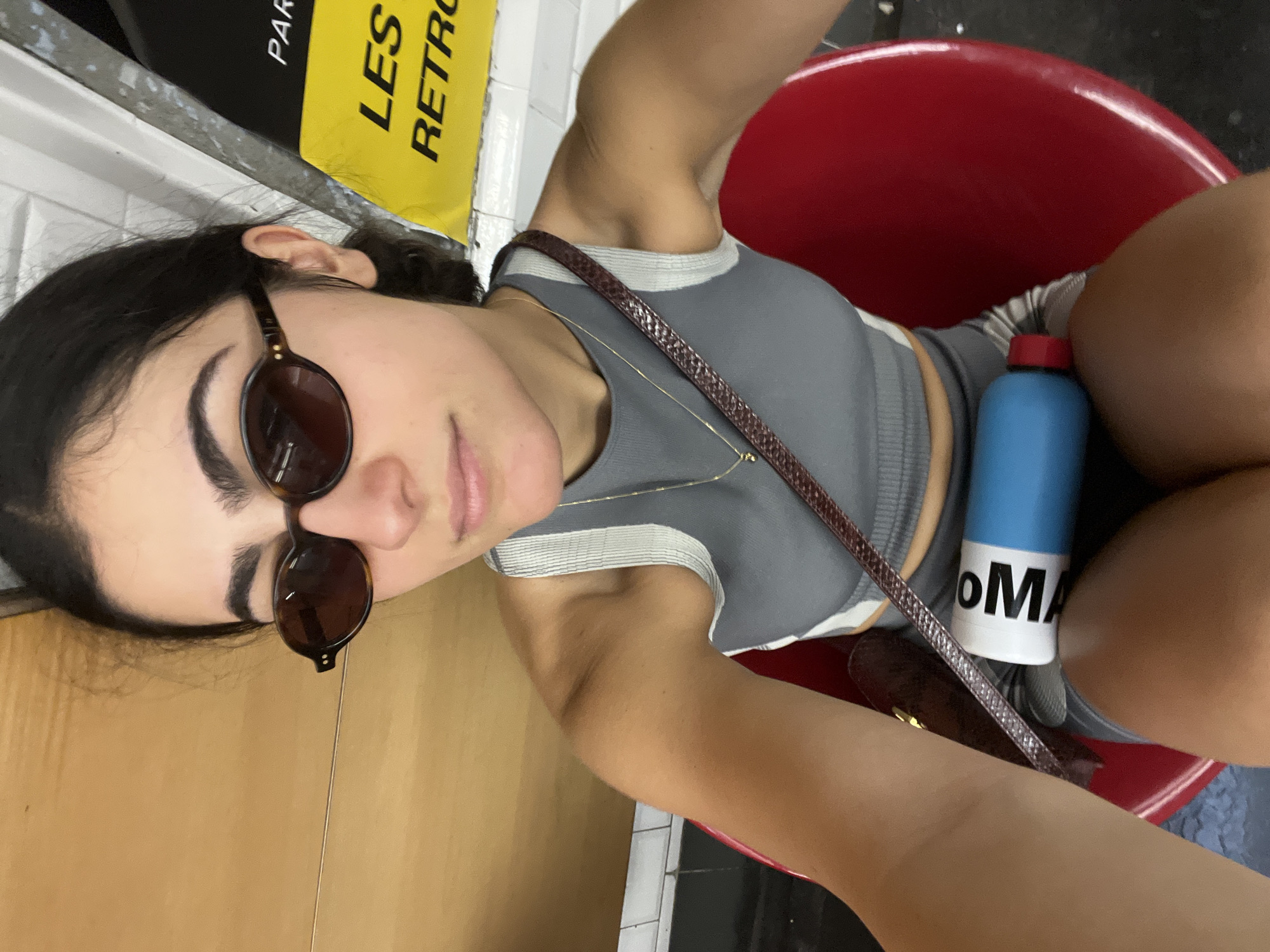 A woman is seated in a red plastic chair in a Paris metro station, wearing sunglasses, a gray cropped top, and shorts. She is holding a blue and white water bottle. A brown purse rests on her lap. Behind her, a yellow sign reads "LES RETRO." The floor is tiled, and a wooden bench is visible on the left side of the frame.