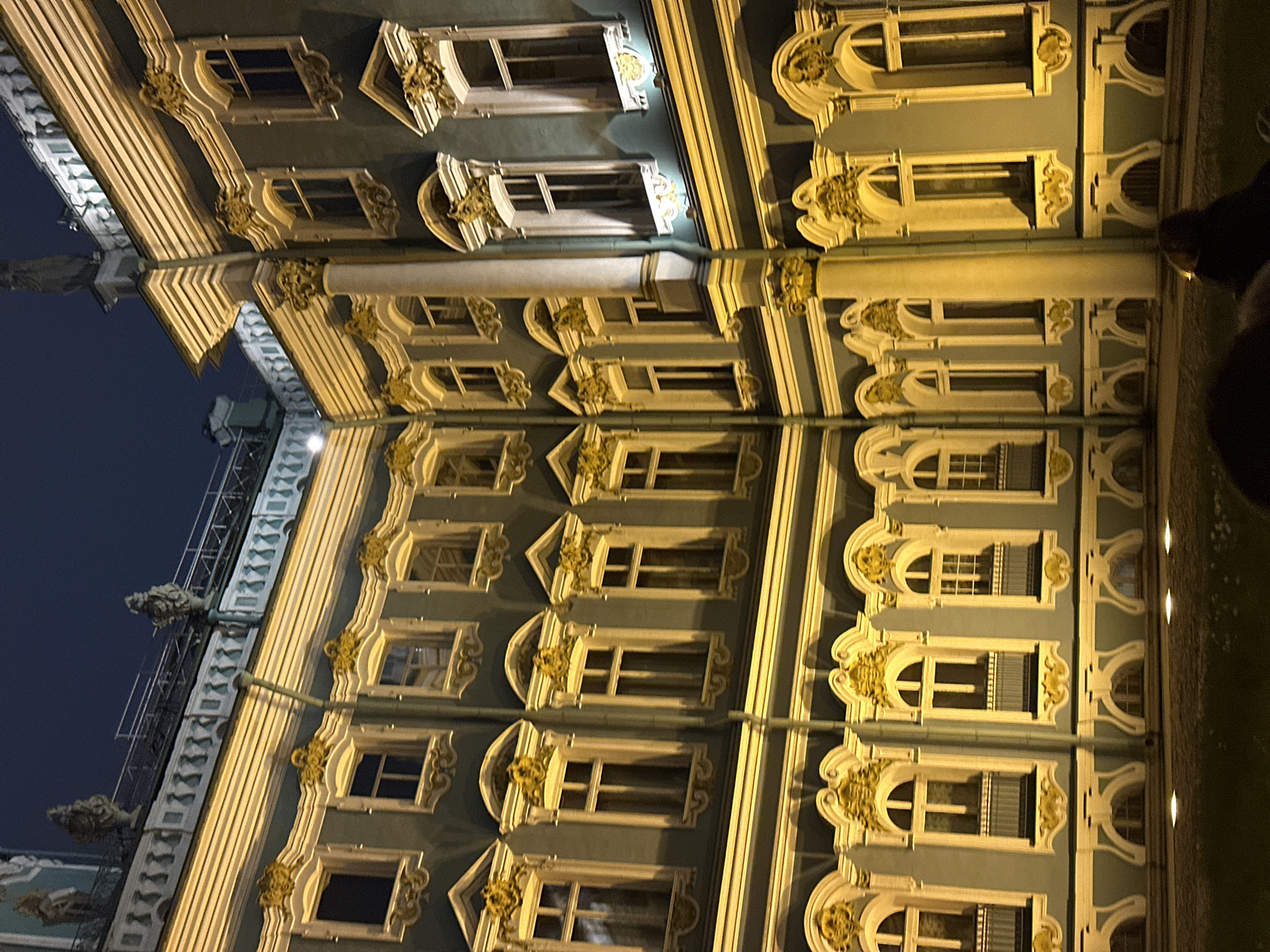 The image shows a building facade in Saint Petersburg, Russia, likely captured at night. The architecture is ornate with multiple windows and decorative trim. The building is illuminated, highlighting the architectural details. A drain pipe is visible, vertically traversing the facade. Scaffolding is visible on the left side of the building.