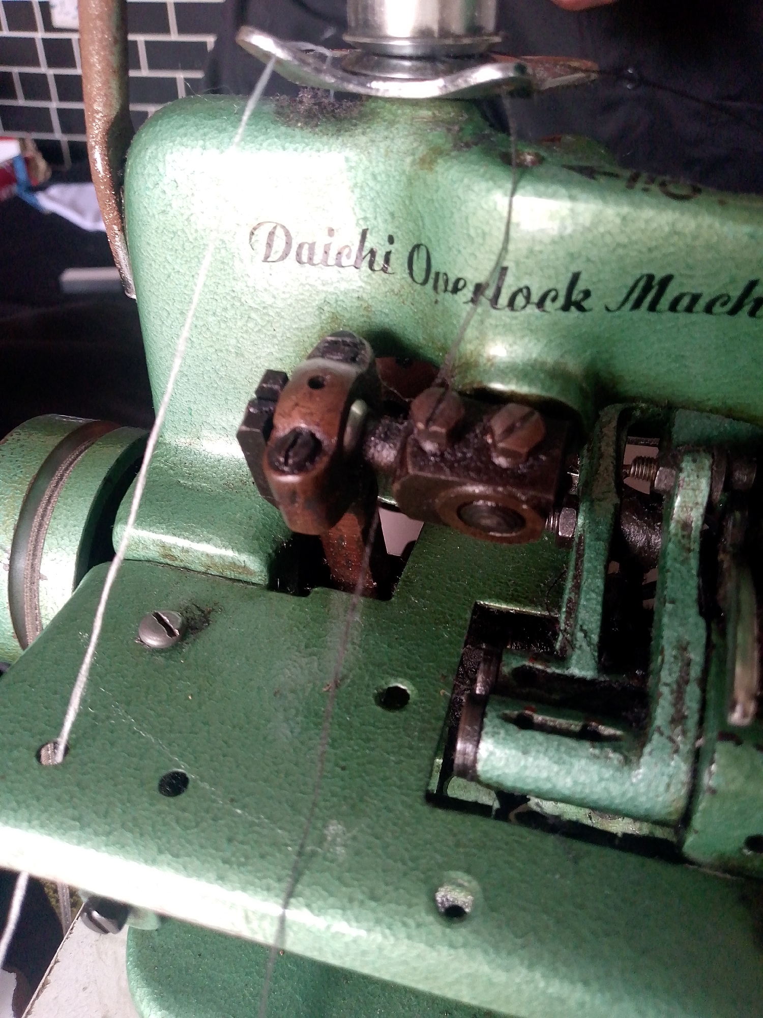 The image shows a close-up of a vintage green Daichi Overlock Machine. The machine appears to be in use, with white thread running through its mechanism. The metal parts of the machine show signs of wear and some oil or grease. The background is slightly out of focus, revealing a brick wall and other indistinct objects, suggesting an indoor setting. There are no people visible in the image. The scene is a static depiction of a sewing machine. The location of Jauro Yinu, Nigeria is not discernible from the visual information provided in the image.