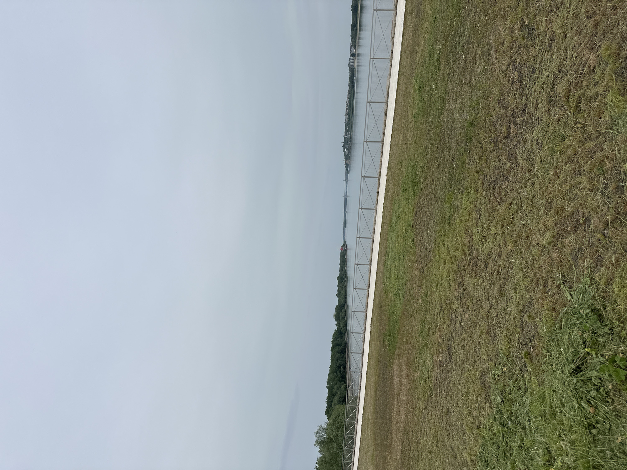The image depicts an outdoor scene along the Danube River in Vidin, Bulgaria. A metal fence runs parallel to the riverbank, separating a grassy area from the water. The river, with visible reflections, stretches towards the horizon under an overcast sky. Across the river, a blurred coastline is visible with hints of buildings and vegetation. The grassy area shows varying shades of green and brown, suggesting different stages of growth or dryness. The perspective suggests the photo was taken from a slightly elevated position.