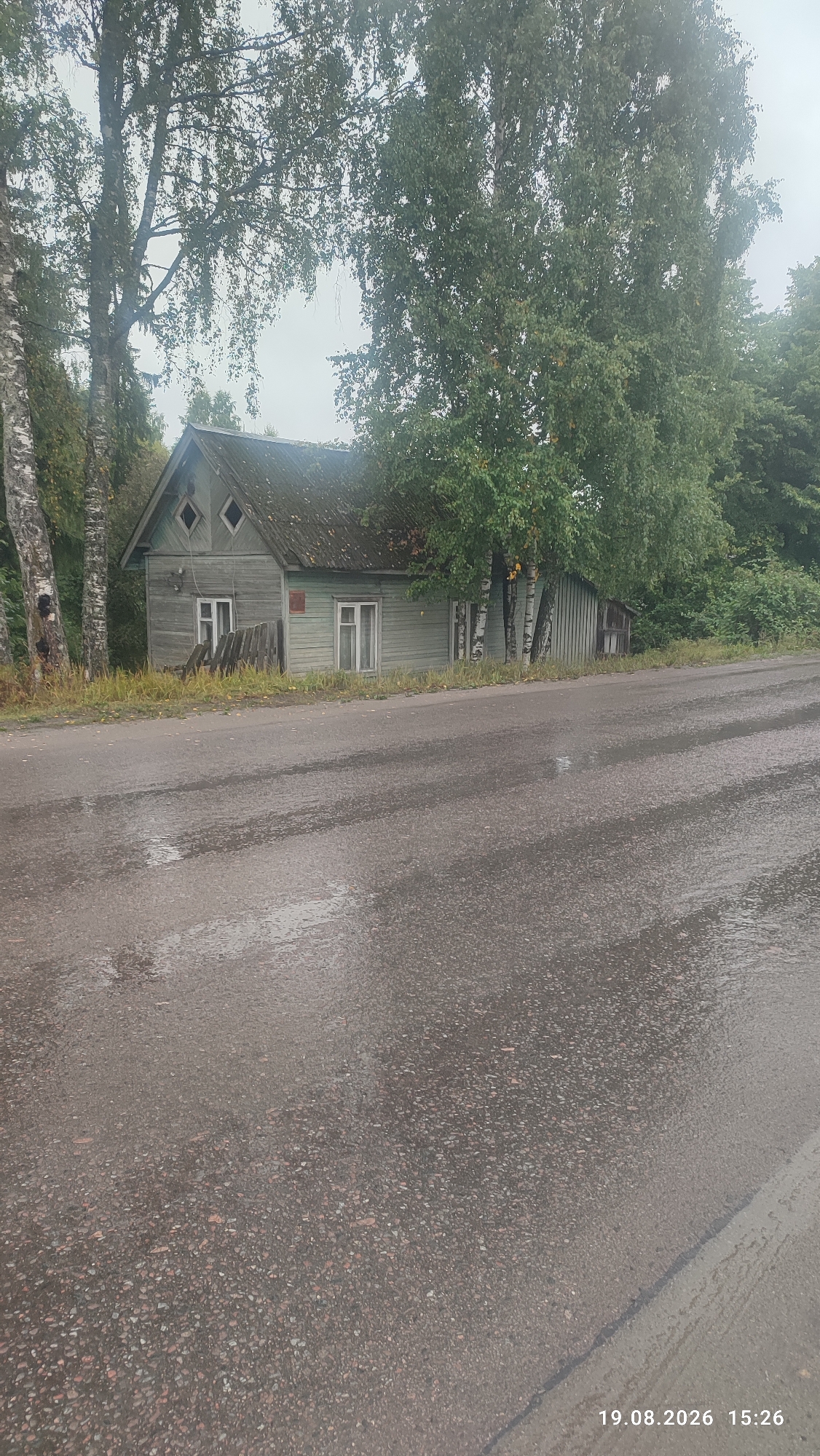 The image captures a weathered, wooden house located in the city of Suoyarvi, Russia. The house, painted a faded green, has a dark, sloping roof. Two diamond-shaped windows are visible on the upper part of the facade, beneath the roofline. White-framed windows are present on the main level. In front of the house, a rustic wooden fence stands partially dismantled. The house is surrounded by trees, with tall birch trees with distinctive white bark framing the left side and a dense canopy of green leaves covering the roofline. The ground in front of the house is covered in dry grass and fallen leaves. A paved road, wet and reflecting the overcast sky, occupies the foreground, with puddles of water indicating recent rainfall. The overall atmosphere is somber and slightly melancholic, suggesting a damp, cloudy day. The timestamp in the lower right corner indicates the image was taken on August 19, 2026, at 15:26.