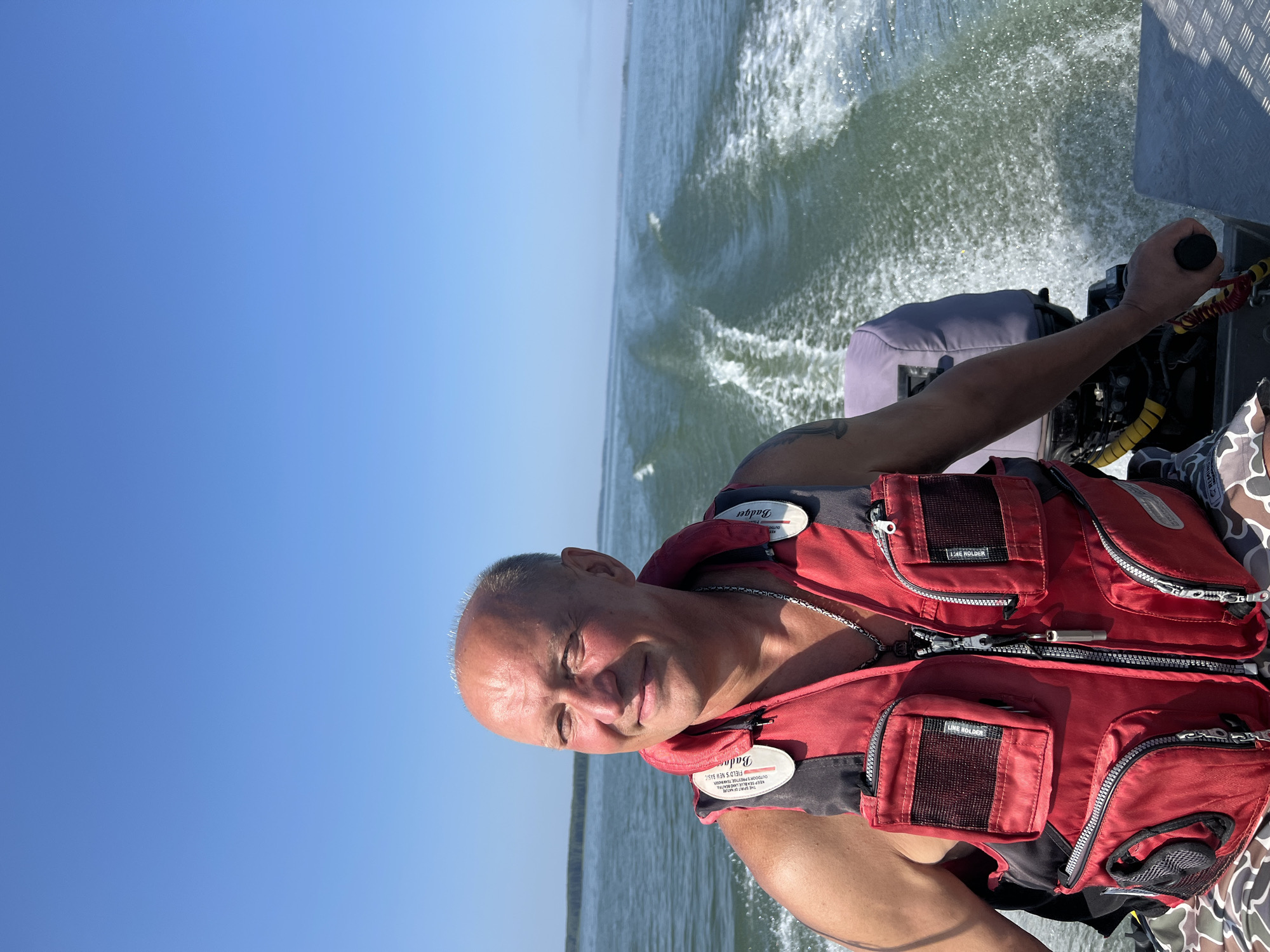 A man, bald with fair skin, is operating a motorboat on a clear day in Beregovoe, Russia. He is wearing a red life vest over a camouflage shirt. The life vest has several zippered pockets and white tags with writing on them. He is looking to his left and smiling. The boat is creating a wake behind it as it moves through the water. The background shows a body of water with a treeline on the horizon under a bright blue sky.