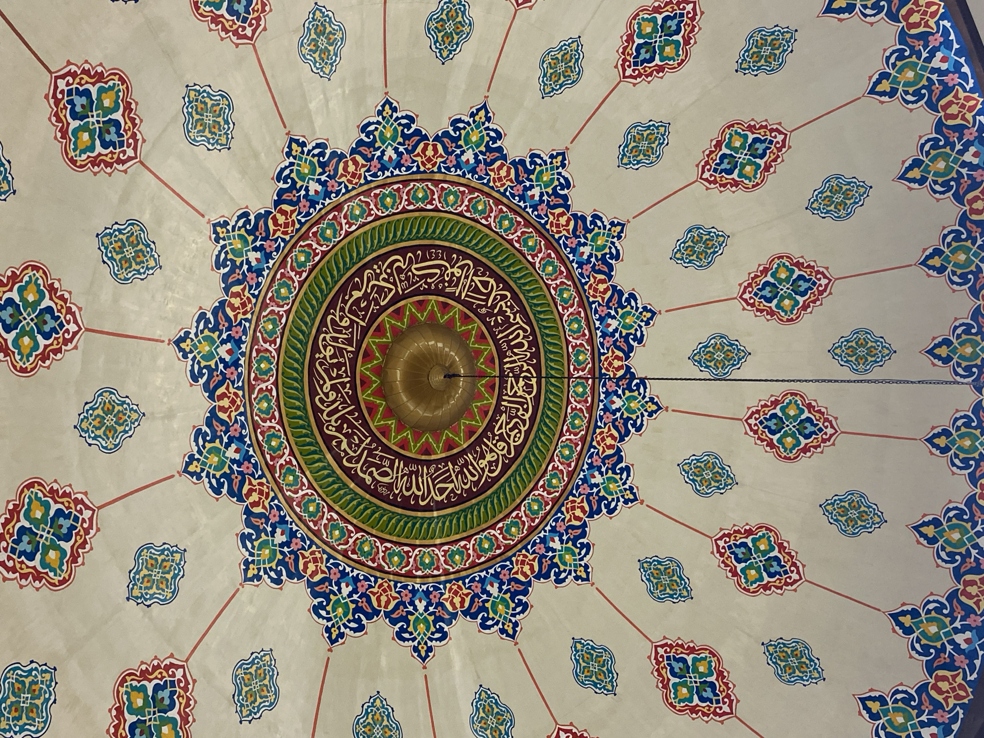 The image shows a close-up, high-angle view of the intricately decorated ceiling of the Islamic Center Mosque in Lhokseumawe, Indonesia. The ceiling features a central medallion with Arabic calligraphy in gold, surrounded by concentric rings of vibrant colors including deep red, green, blue, and gold. This central design radiates outwards with elaborate floral and geometric patterns. Smaller, repeating decorative motifs in similar colors are distributed across the beige ceiling, connected by thin red lines. A golden, dome-shaped fixture, likely a lamp, is suspended from the center of the ceiling. The overall impression is one of ornate Islamic architectural design.