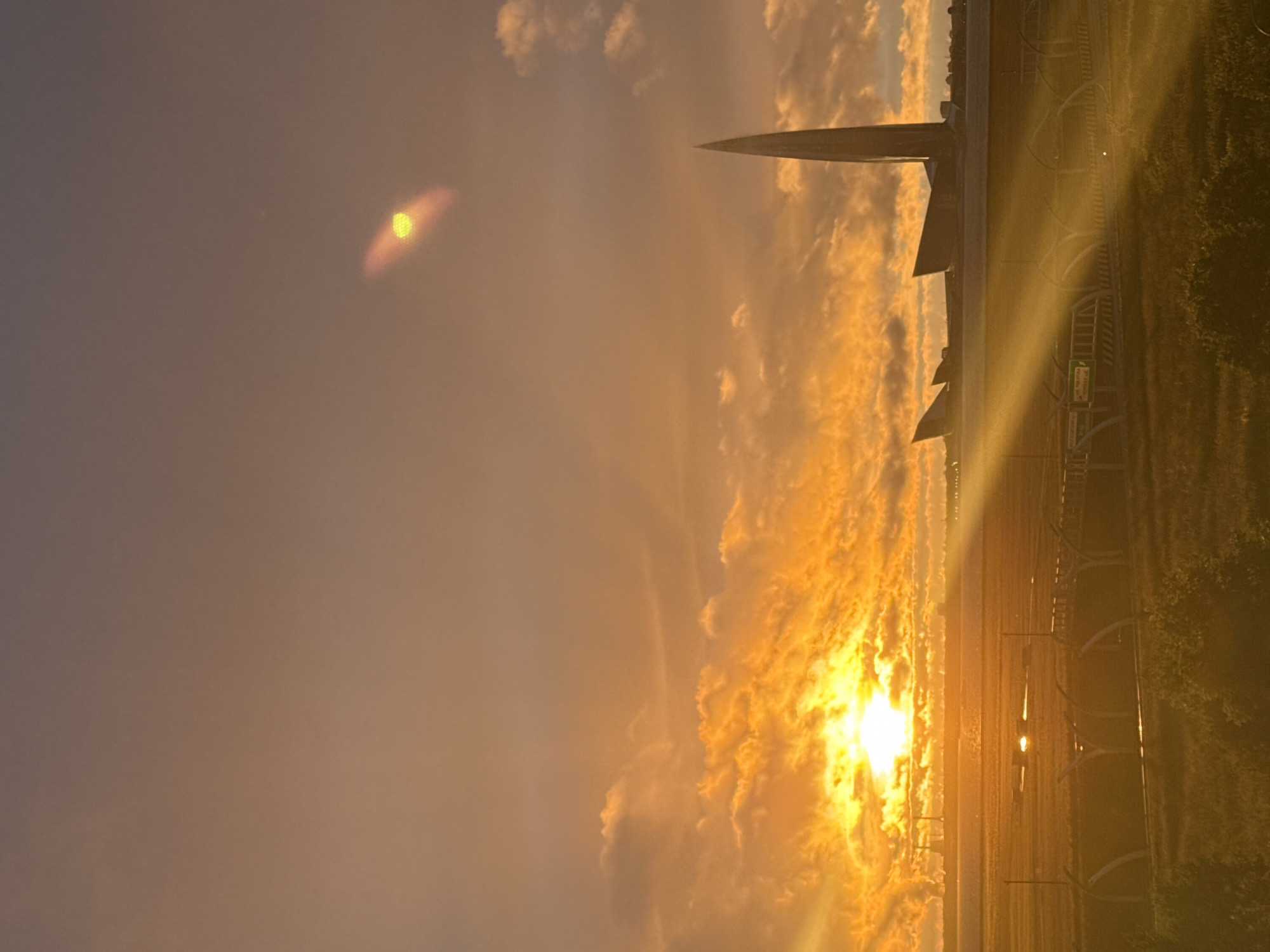 This is a vertical, eye-level, outdoor photograph taken at sunset. The sky is filled with dramatic, fiery orange and yellow clouds, with the sun casting a bright glow. In the foreground, a grassy field is visible with a road and fencing. In the middle ground, there is a large body of water reflecting the sunset. In the background, a modern, sleek skyscraper with a pointed top dominates the skyline, identified as the Lakhta Center in Saint Petersburg, Russia. Two smaller, wing-like structures are also visible near the skyscraper. The overall impression is one of a dramatic and somewhat futuristic urban landscape bathed in the warm light of a setting sun.