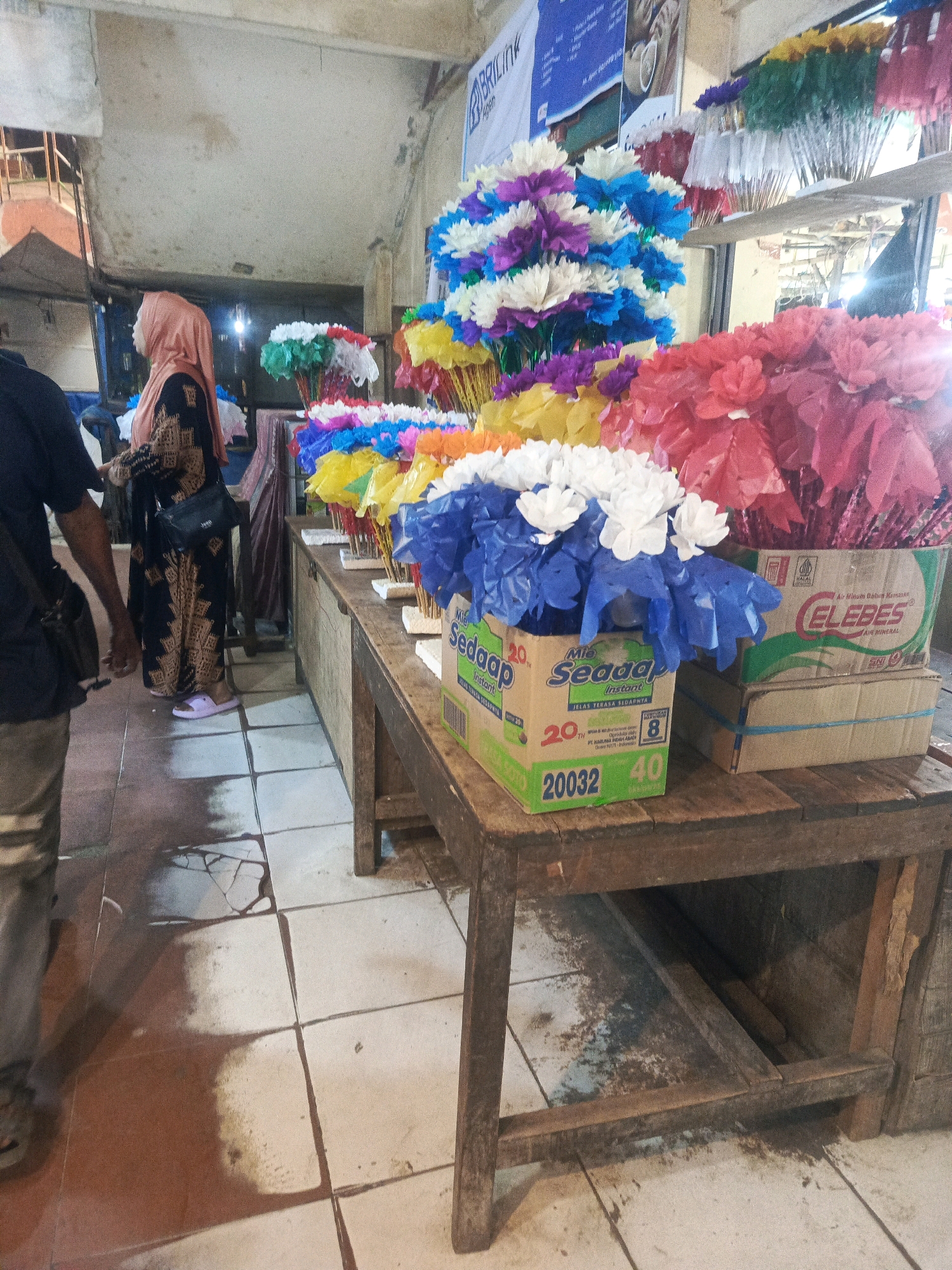 The image captures a market stall in Kendari, Indonesia, likely a souvenir or craft shop. The foreground features a rustic wooden table laden with various colorful artificial flowers, predominantly in shades of red, pink, blue, white, and purple. These are arranged in bundles, some contained within cardboard boxes, one of which bears the "Mie Sedaap" brand and another with "ELEBES" and "AIR MINERAL" text. Behind the table, a woman wearing a peach-colored hijab and a patterned black and gold dress is partially visible, seemingly interacting with a vendor or browsing. To the left, a person in a dark blue shirt and tan pants is also present, their back mostly to the camera. The stall is set against a backdrop of a concrete structure with exposed beams and what appears to be additional merchandise, including more flower arrangements and possibly other decorative items, displayed on shelves. The floor is tiled, with some areas appearing wet or stained, suggesting a bustling and potentially humid environment. A promotional banner with blue and white text is affixed to the wall in the upper portion of the frame. The overall scene depicts a vibrant commercial setting within the city.